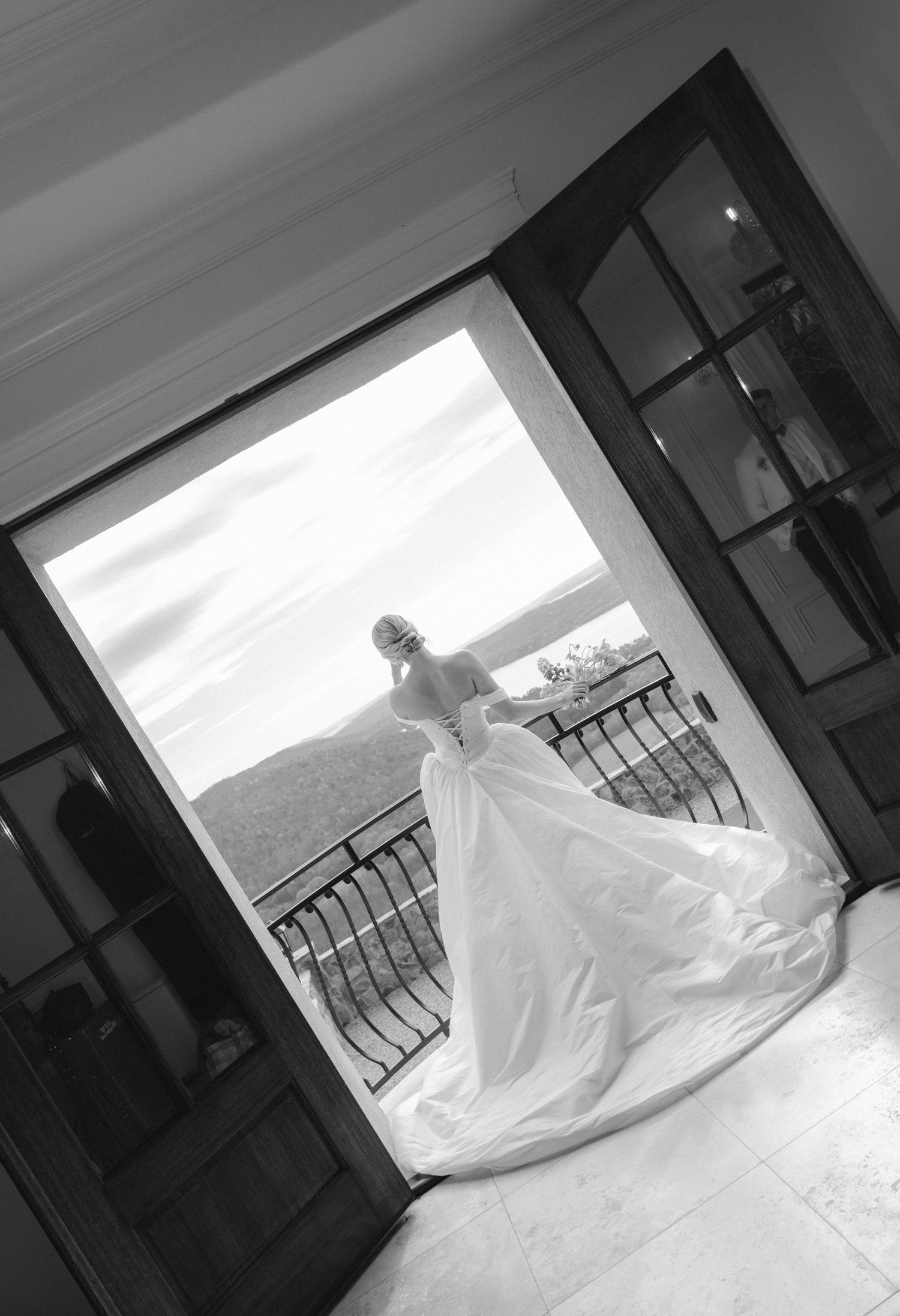 Black and white photo of a bride in a flowing gown standing in open French doors on a balcony with scenic views.