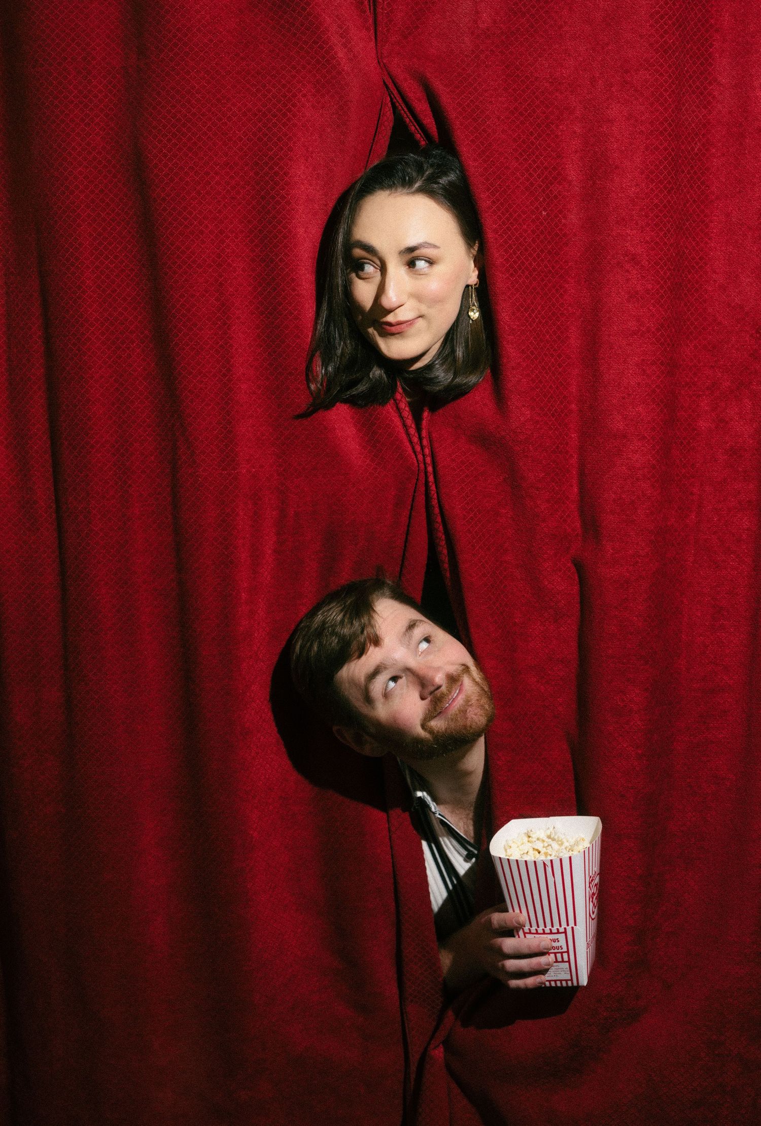 Two people peek through red curtain holes, man holding popcorn bucket, woman above smiling playfully.