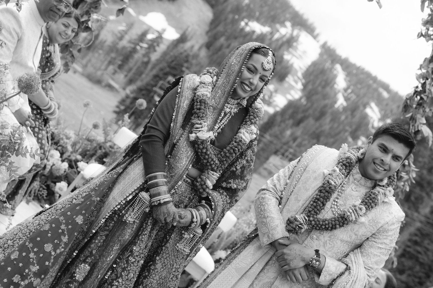 Black and white photo of Indian bride and groom laughing during traditional wedding ceremony outdoors.