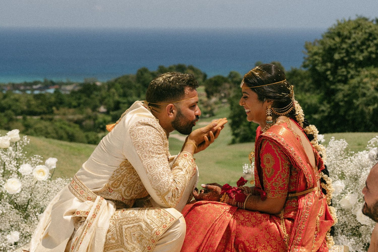 Indian couple in traditional wedding attire sharing a romantic moment outdoors with ocean views in the background.