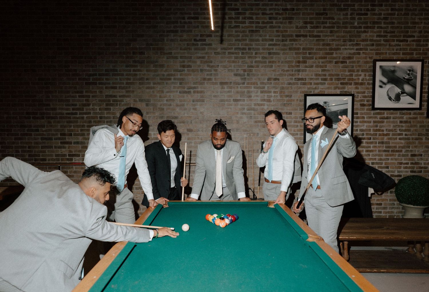 Group of seven men in white suits playing pool on a green billiard table in a brick-walled room.