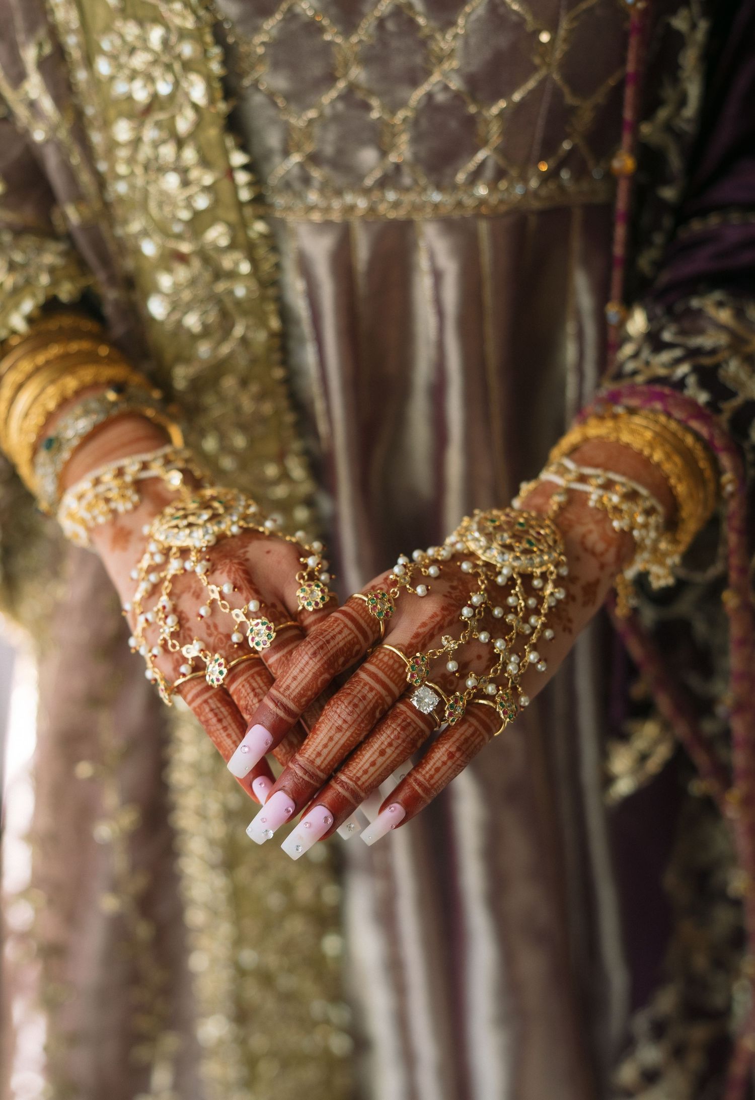 Close-up of South Asian bride's hands adorned with intricate henna, gold bangles, rings, and floral jewelry against embroidered outfit.