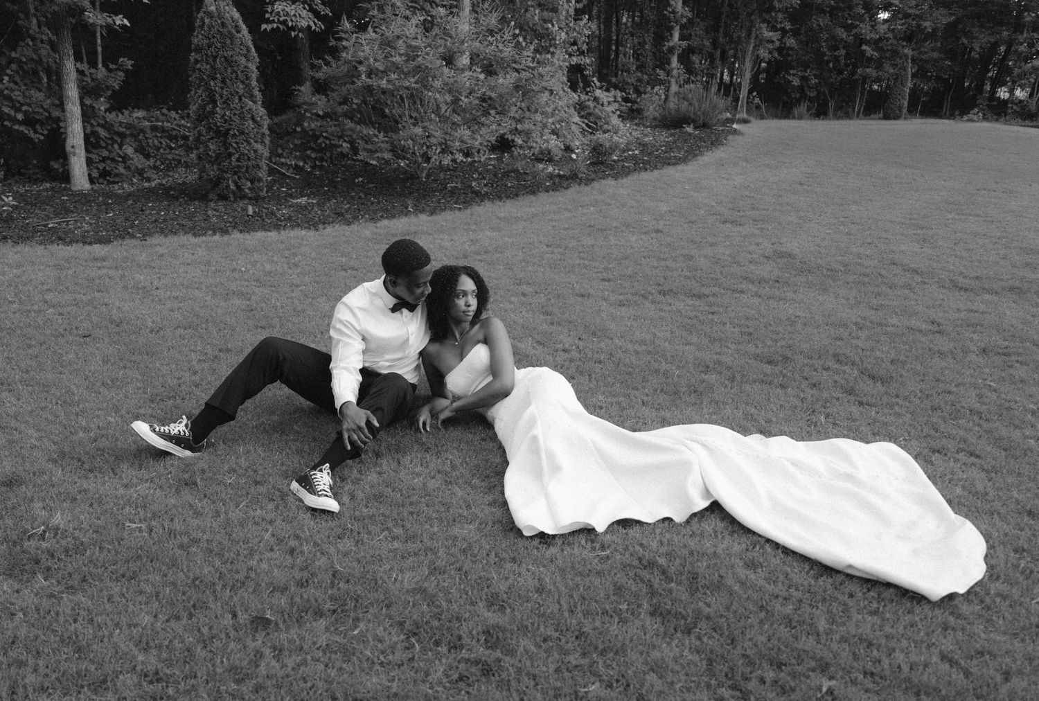 Black and white photo of a bride in white gown and groom sitting together on lush green grass outdoors.
