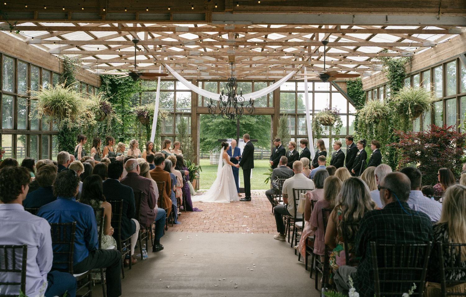 Elegant greenhouse wedding ceremony with couple at altar, guests seated in rows, draped fabric ceiling, and lush greenery backdrop.