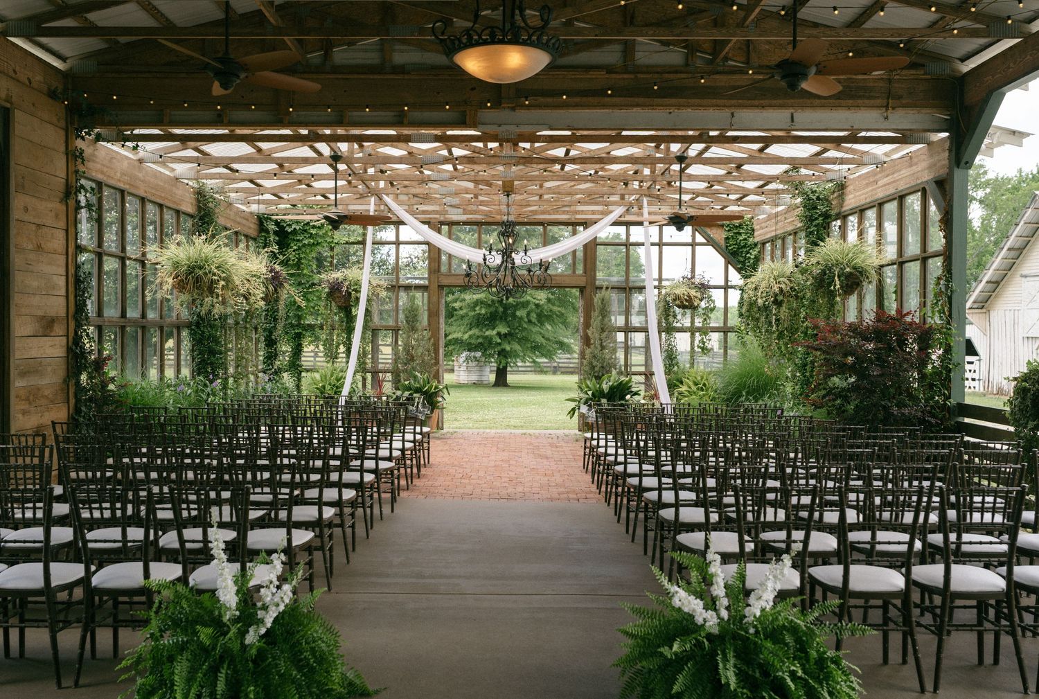 Elegant indoor wedding ceremony venue with lush greenery, rows of dark chairs, and a pergola ceiling draped with white fabric.