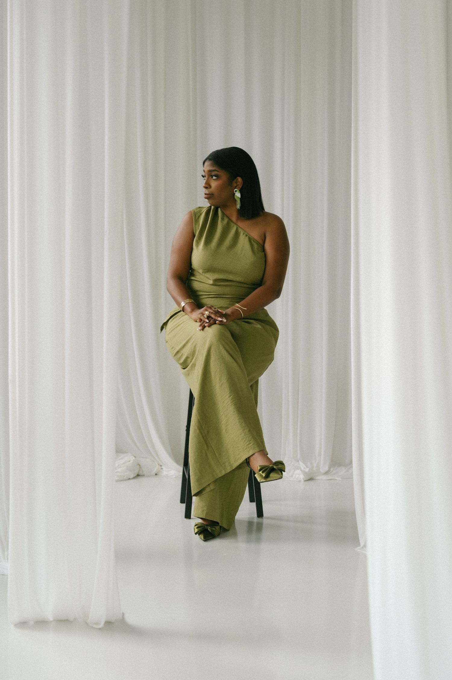 Woman in elegant olive green one-shoulder maxi dress sits gracefully against white curtain backdrop.