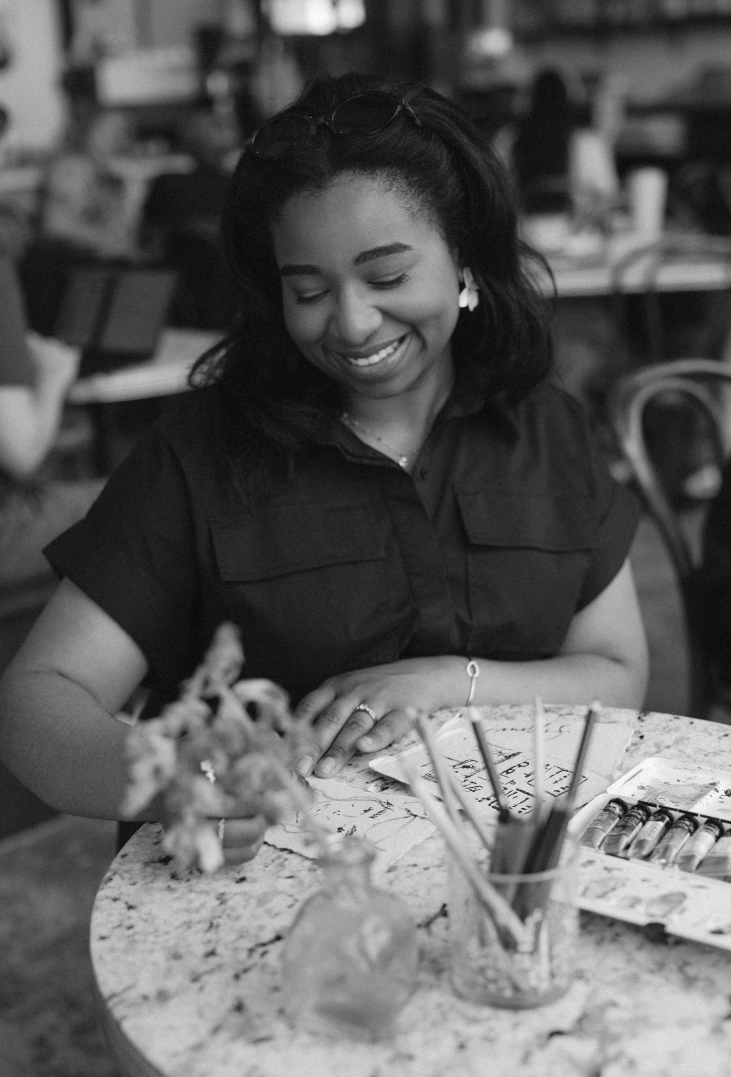 Black and white photo of a smiling woman sitting at a table with paintbrushes and art supplies.