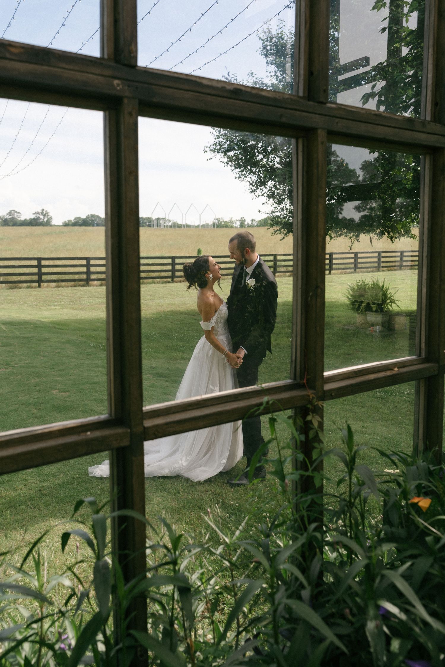 A bride and groom share a romantic moment outdoors on a lush green farm, photographed through a rustic wooden window frame.