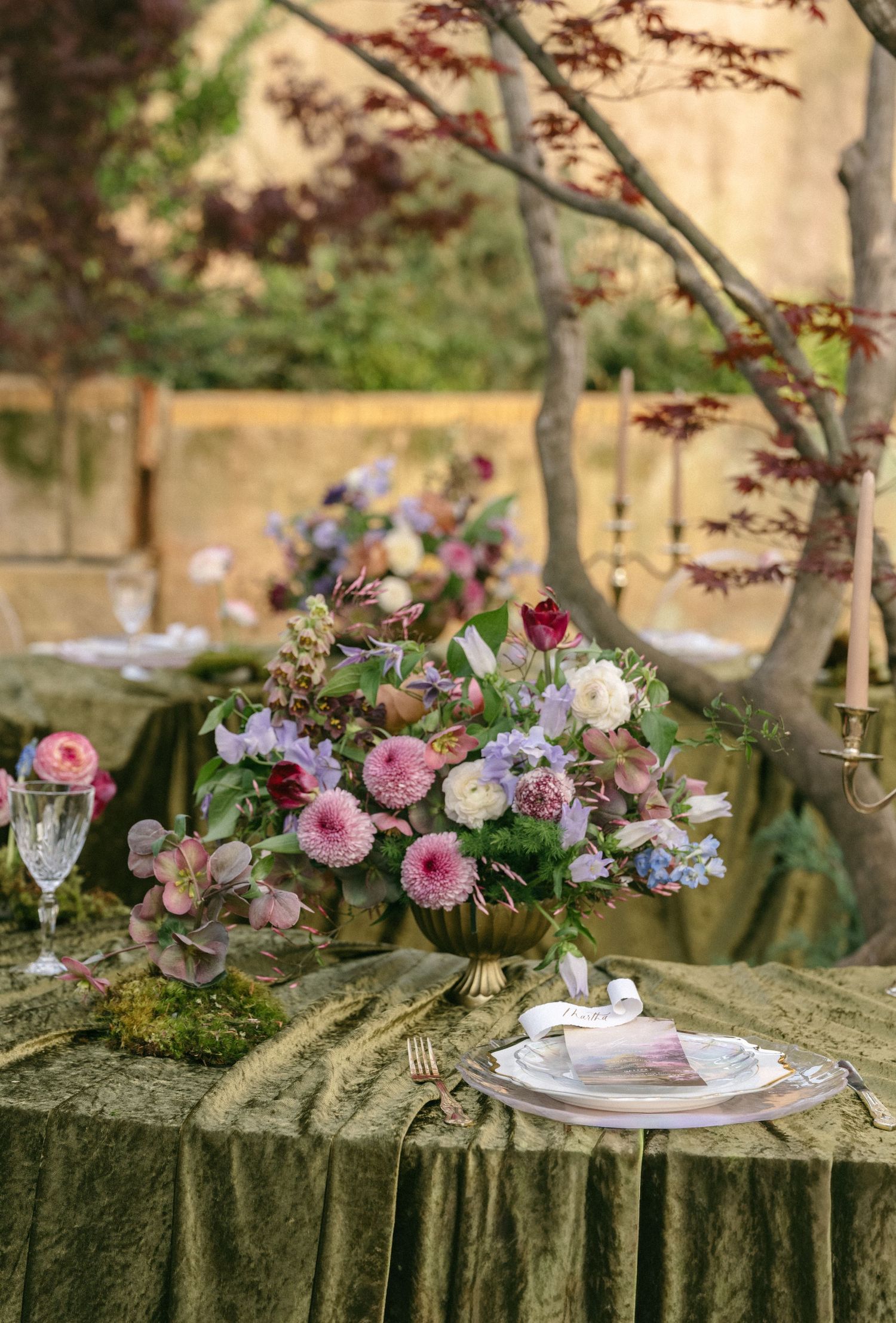 Elegant outdoor wedding tablescape with lush green velvet linens, a gold floral centerpiece of pink and purple blooms, and fine china.