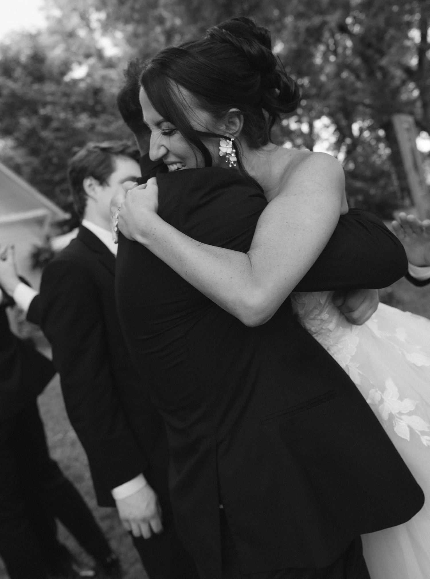 Black and white photo of a bride in a floral gown sharing a joyful hug with a guest in a tuxedo at a wedding.