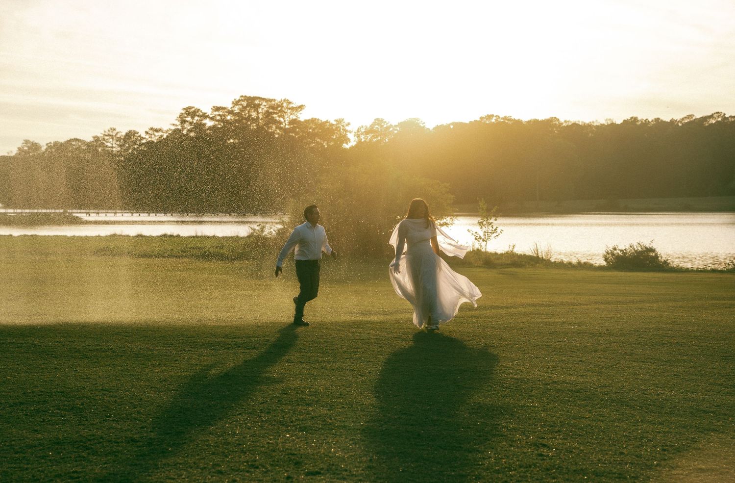 Bride and groom walking hand in hand toward a sunlit lake at golden hour on their wedding day.