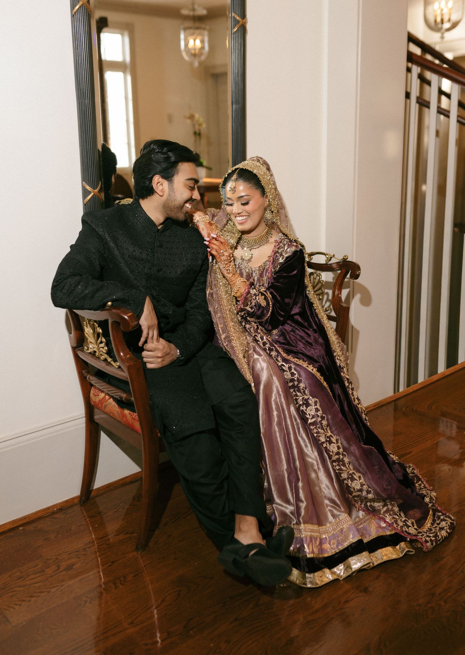 South Asian couple in traditional black and purple wedding attire sitting on a vintage chair in an elegant home.