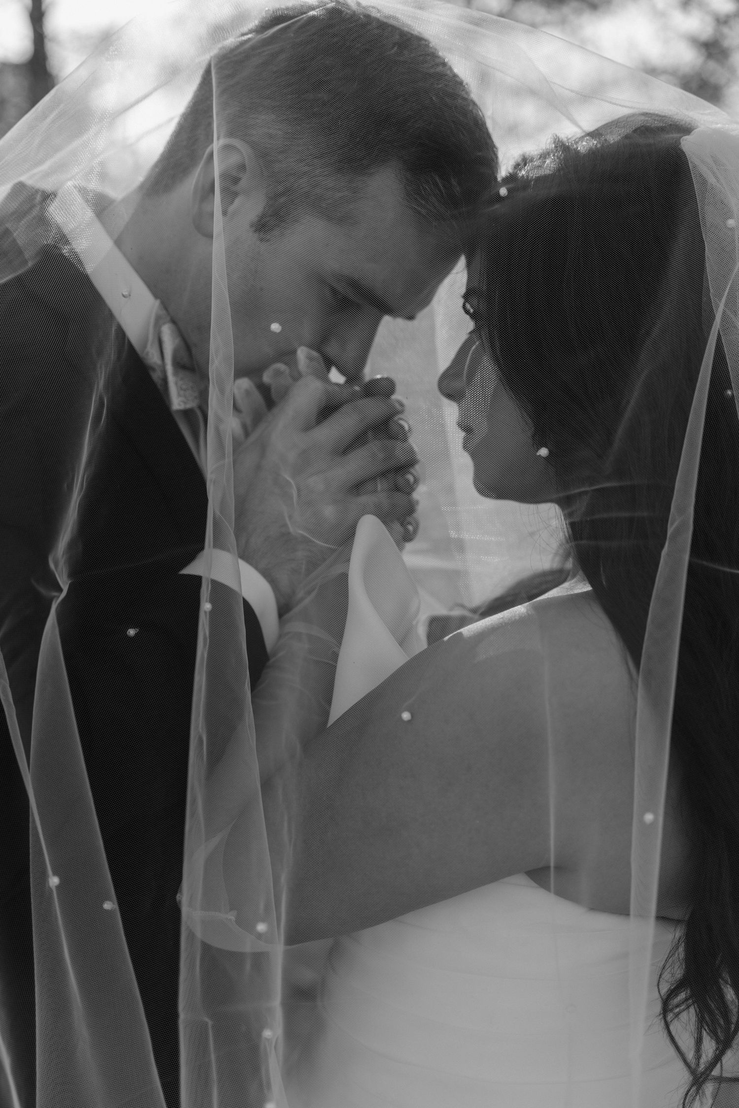 Black and white photo of bride and groom holding hands under wedding veil, foreheads touching in romantic pose.