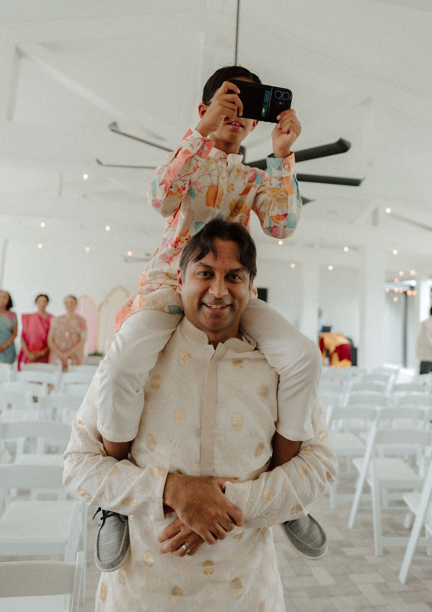 Smiling groom in ivory kurta holds a child on his shoulders taking photos inside a bright wedding venue.