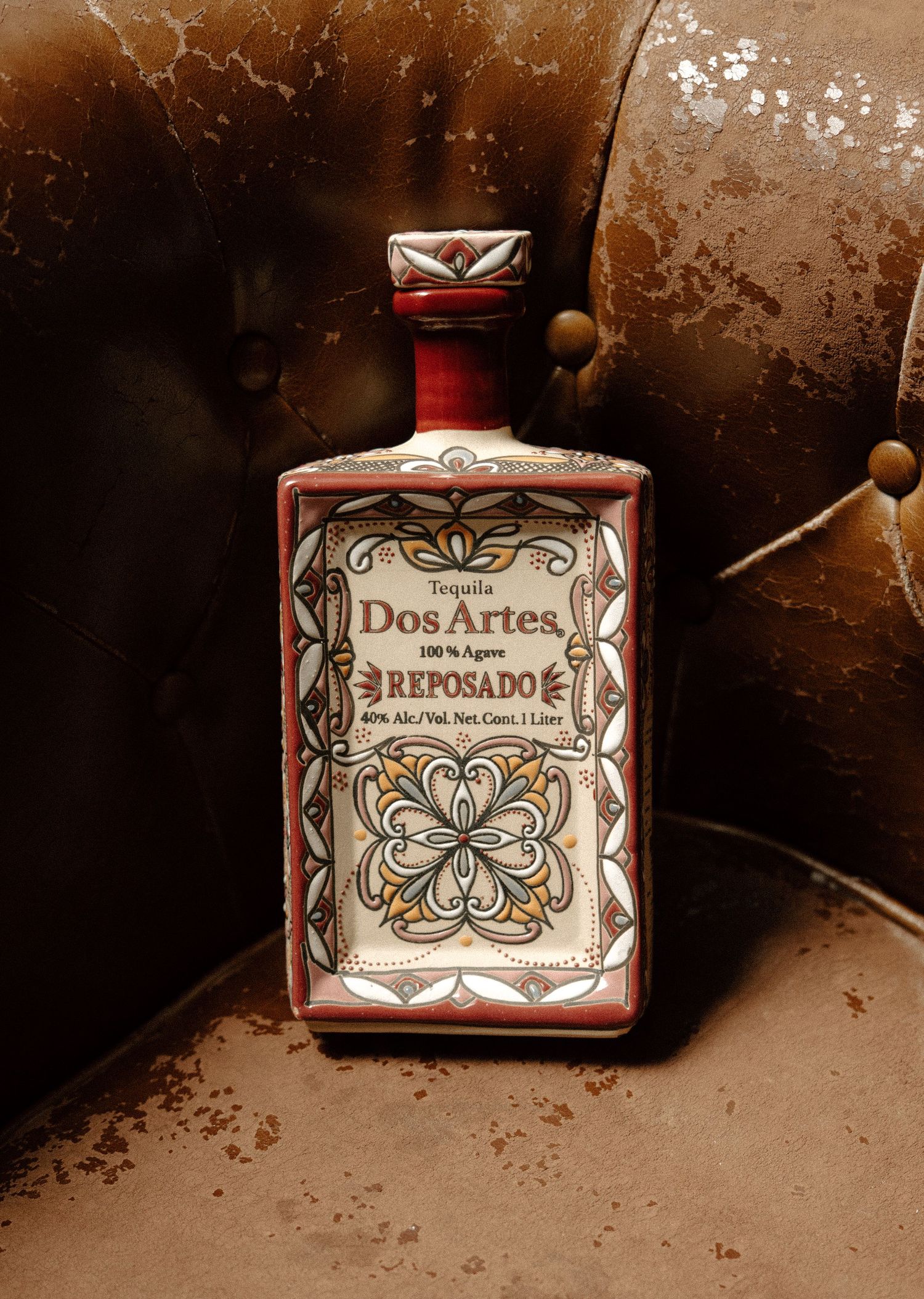 Dos Artes Reposado Tequila 1 liter bottle with ornate floral ceramic design, resting against vintage leather chair.