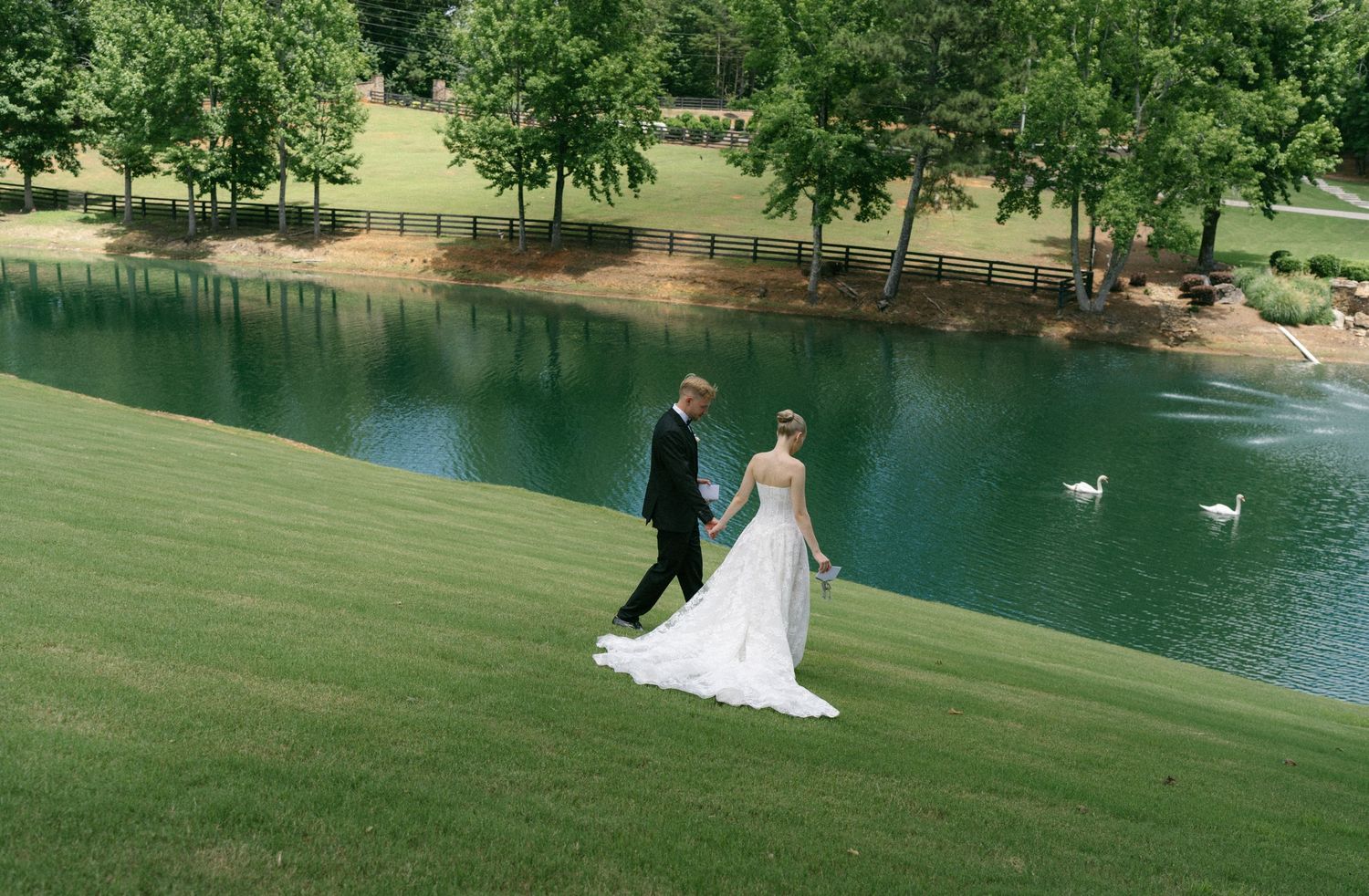 Bride and groom walking hand in hand on lush green lawn beside a serene pond with swans on a sunny wedding day.