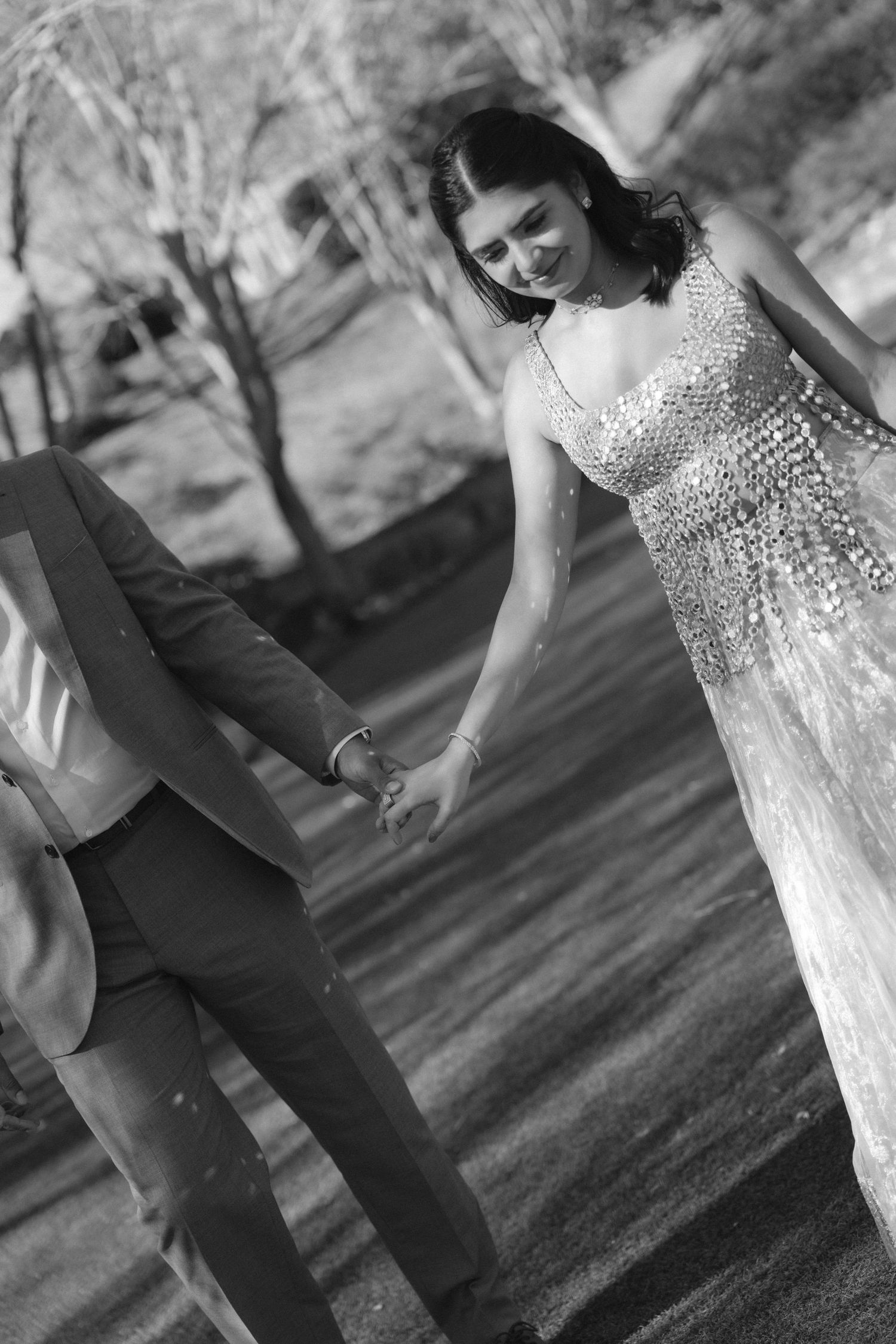 Black and white photo of a couple holding hands outdoors, woman in sequined gown smiling at man in suit.