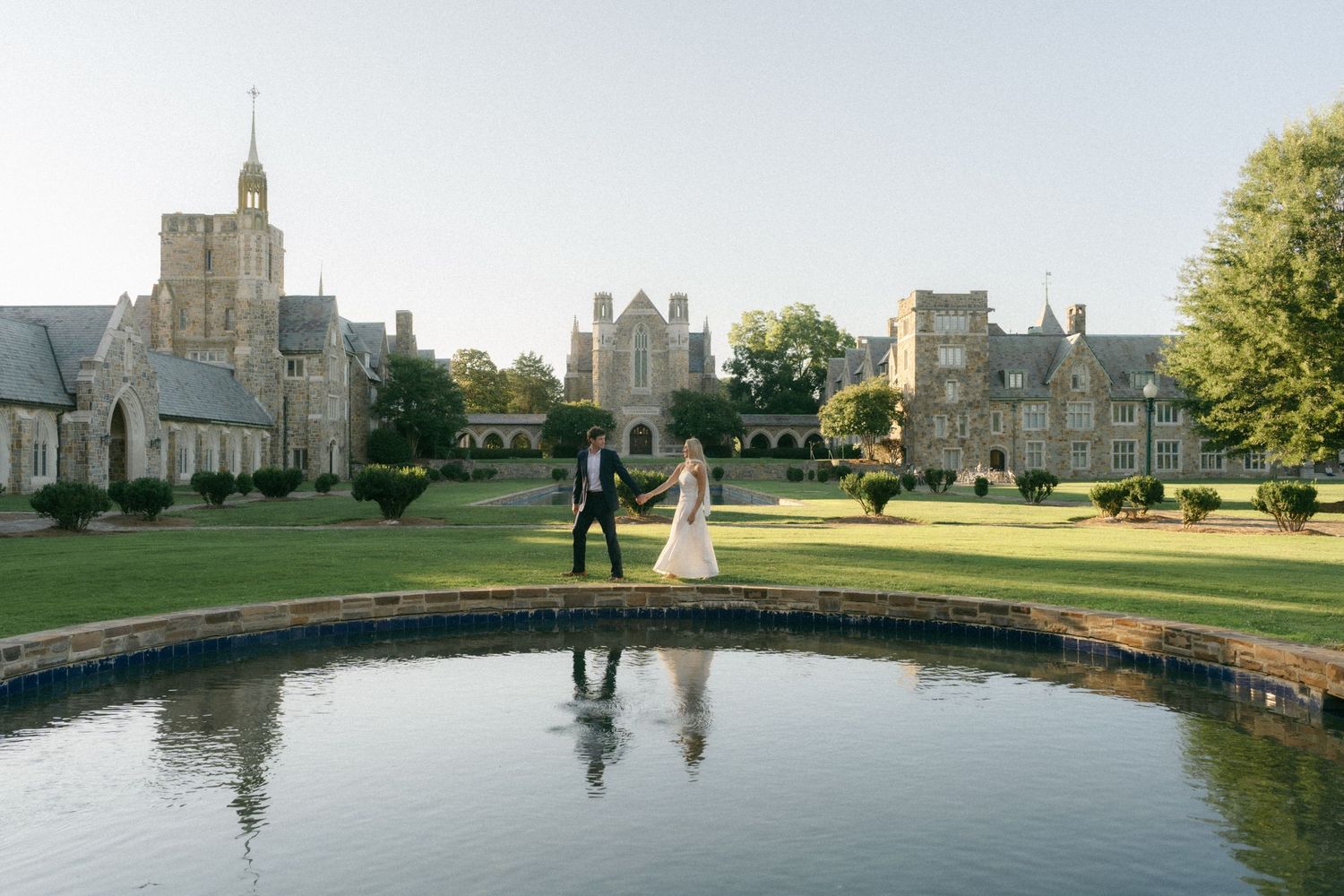 Bride and groom walk hand in hand beside a reflecting pond at an elegant historic estate on a sunny day.