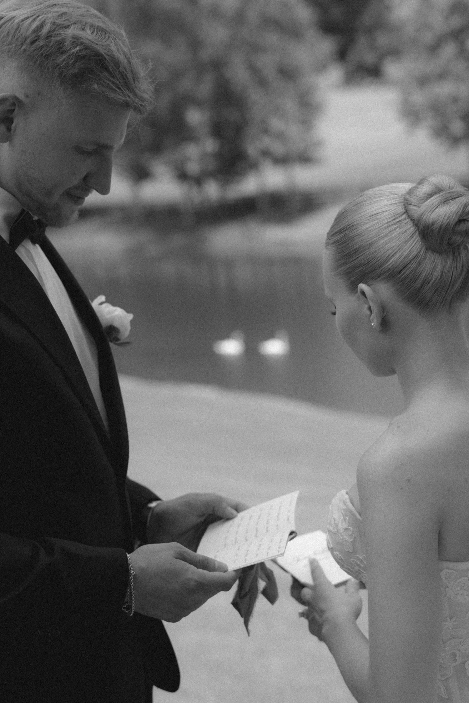 Black and white photo of bride and groom exchanging handwritten vows outdoors near a serene pond.