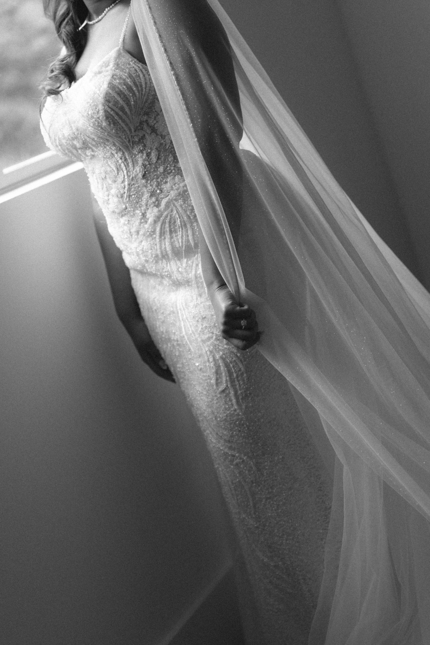 Bride in fitted lace wedding gown with flowing veil, black and white portrait photo.