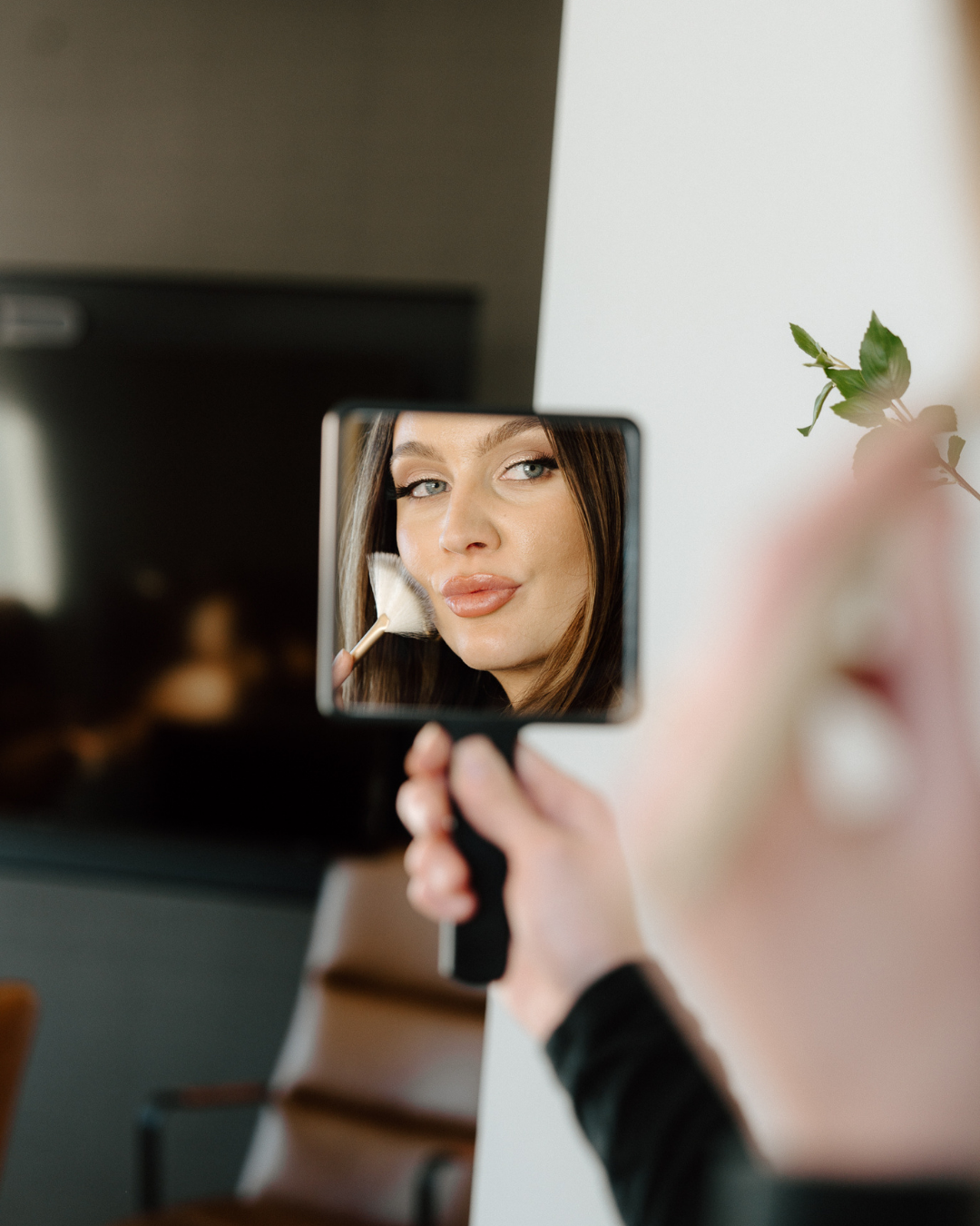 Woman applying makeup with a brush, reflected in a handheld mirror, with a cozy living room in the background.