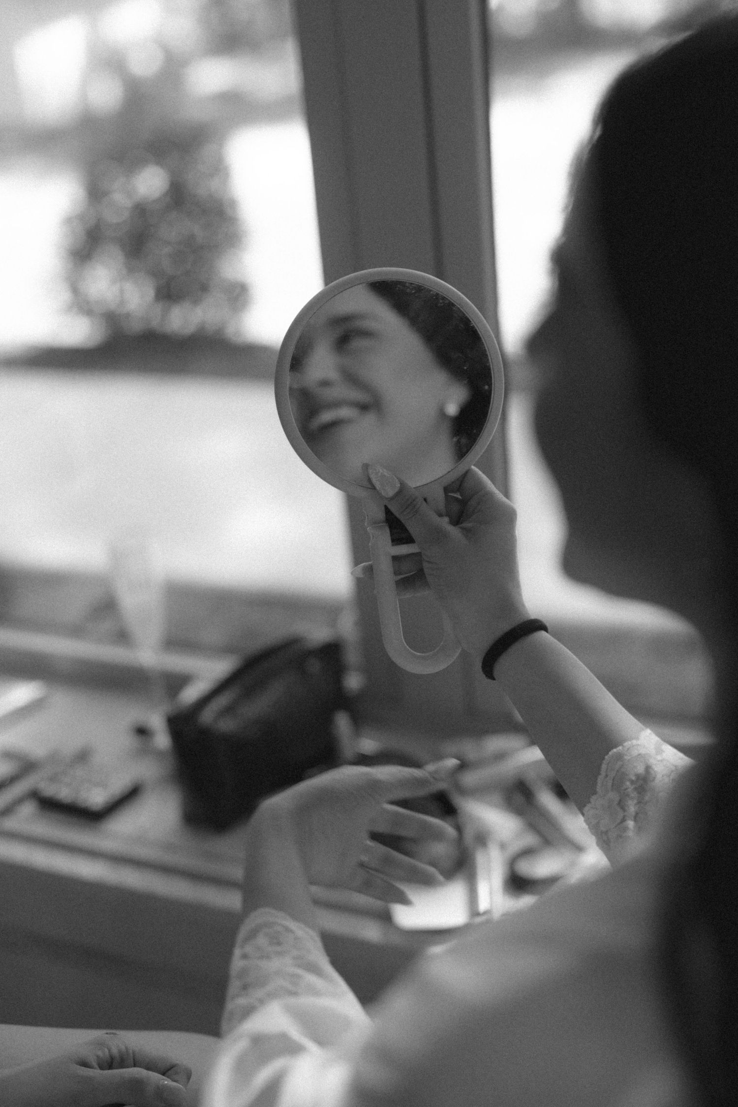 Black and white photo of a smiling bride viewing her reflection in a small handheld mirror during wedding preparations.