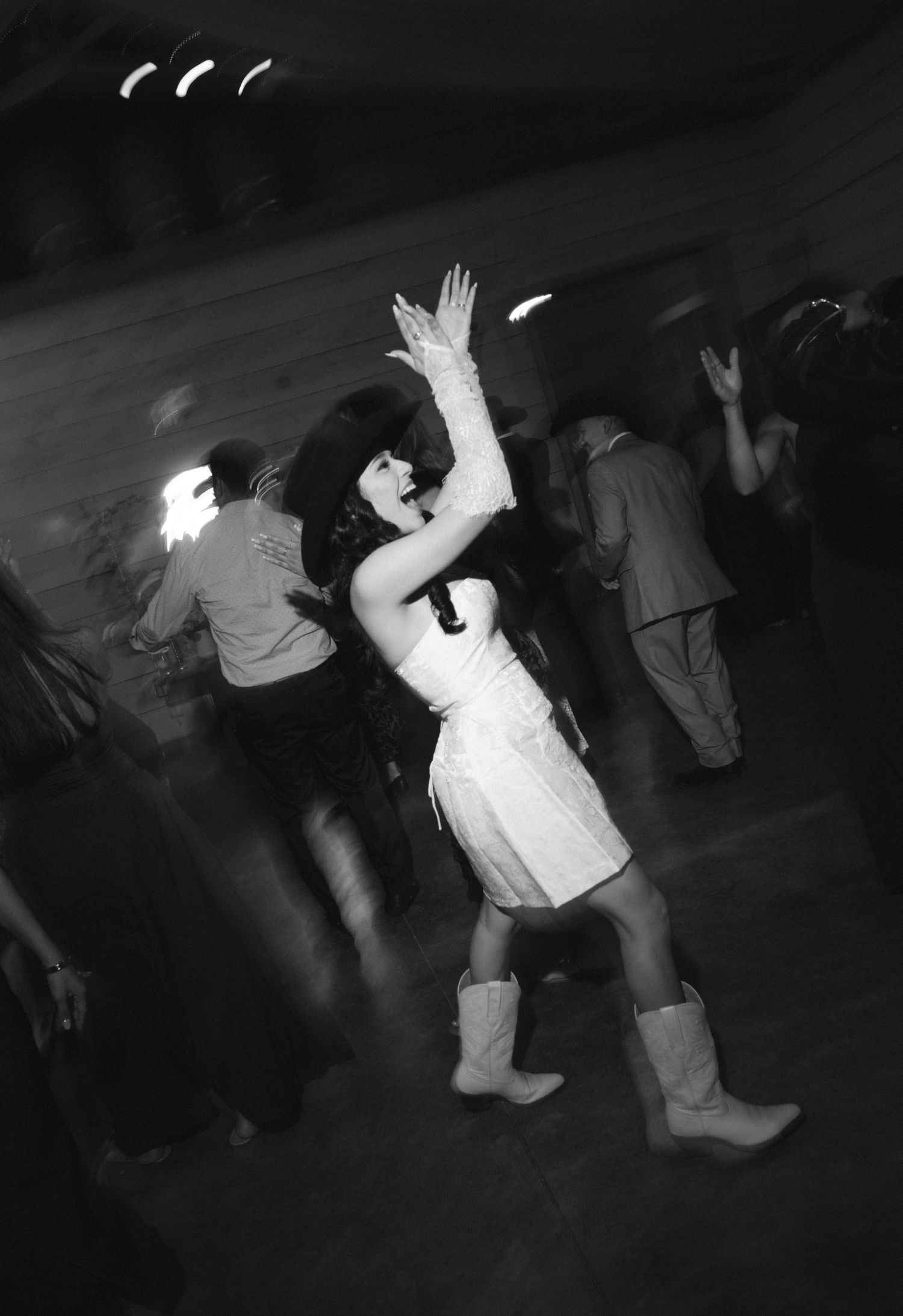 Black and white photo of a woman in a white dress and cowboy boots dancing energetically at a party or wedding reception.