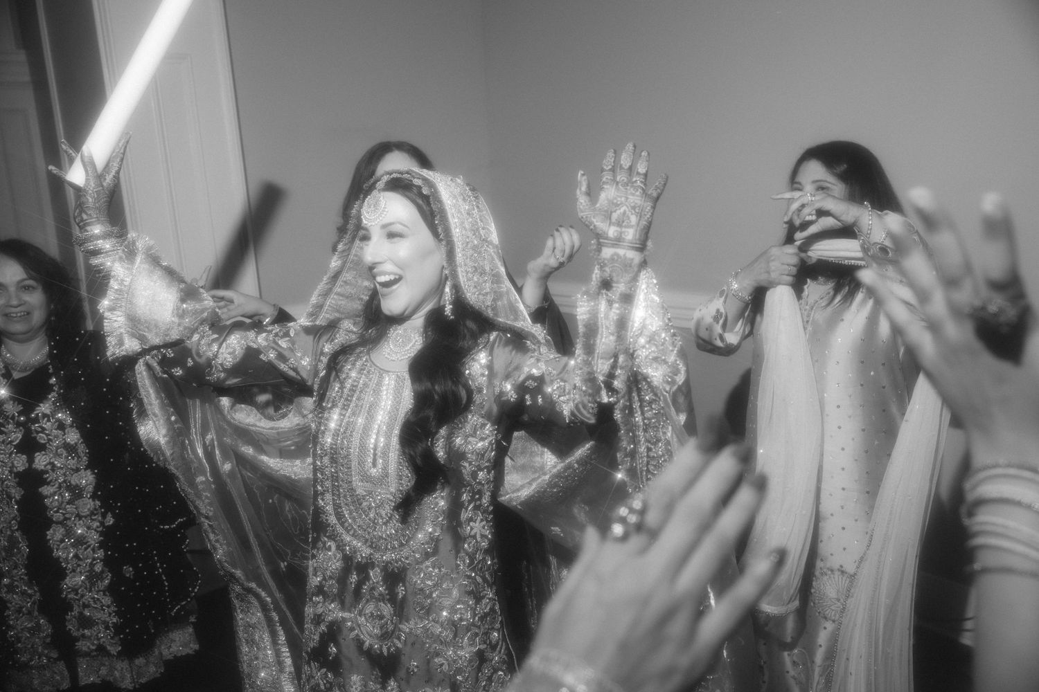 Black and white photo of a bride in traditional South Asian attire dancing joyfully with guests at a wedding celebration.