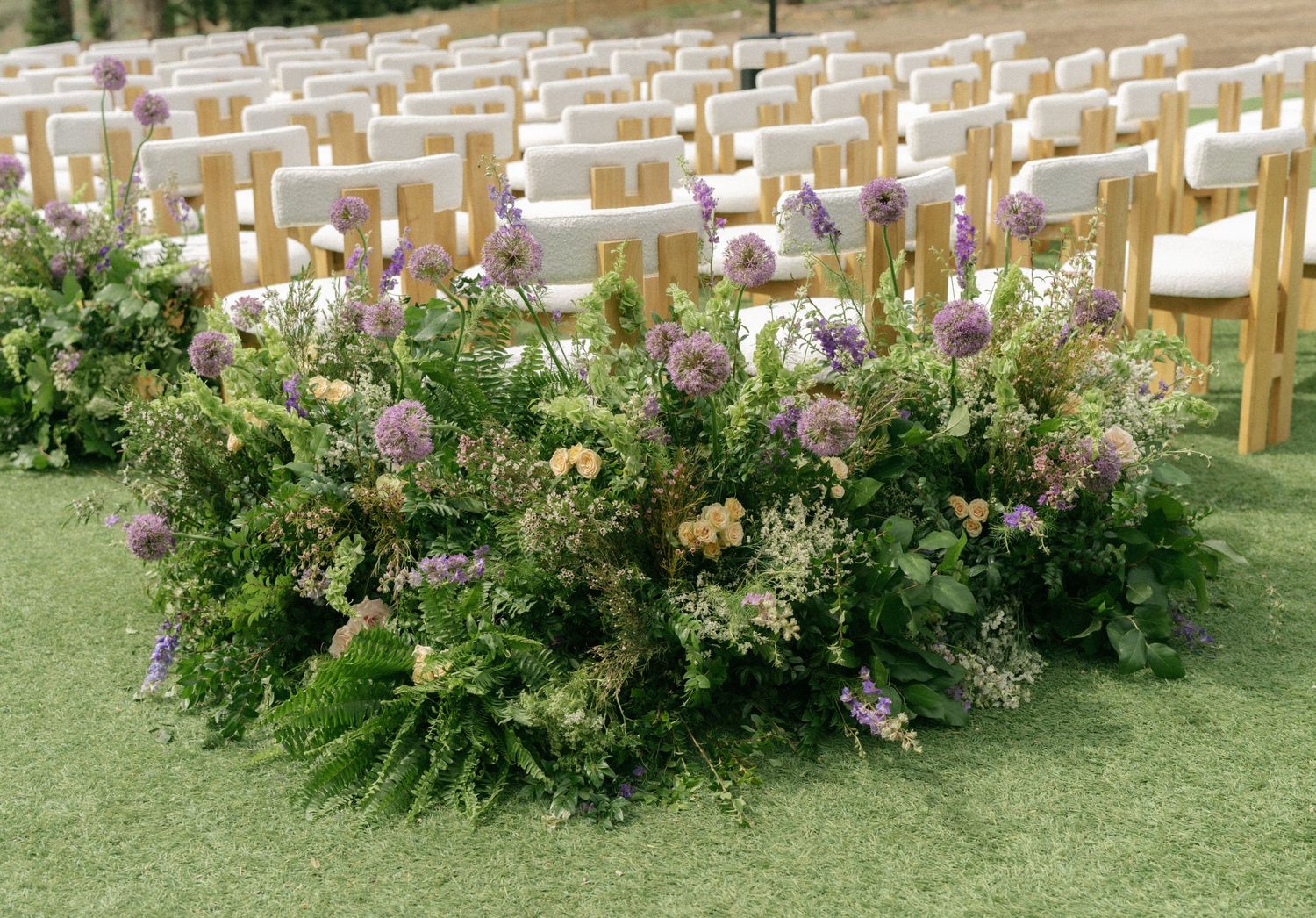 Lush floral arrangement with purple alliums and wildflowers lining outdoor wedding ceremony aisle with white chairs.