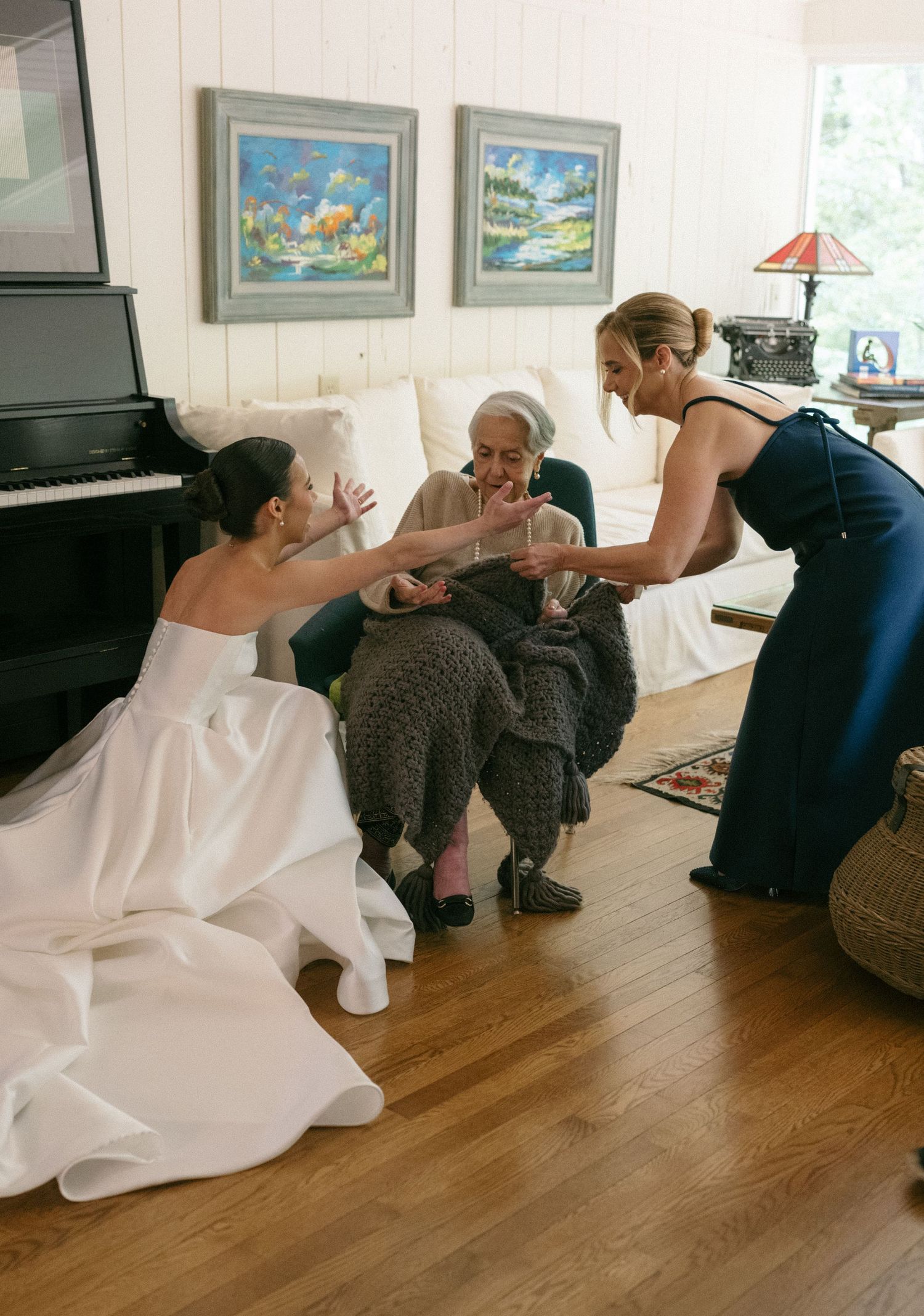 Bride and bridesmaid in navy dress kneeling to show elderly seated woman wedding ring indoors.
