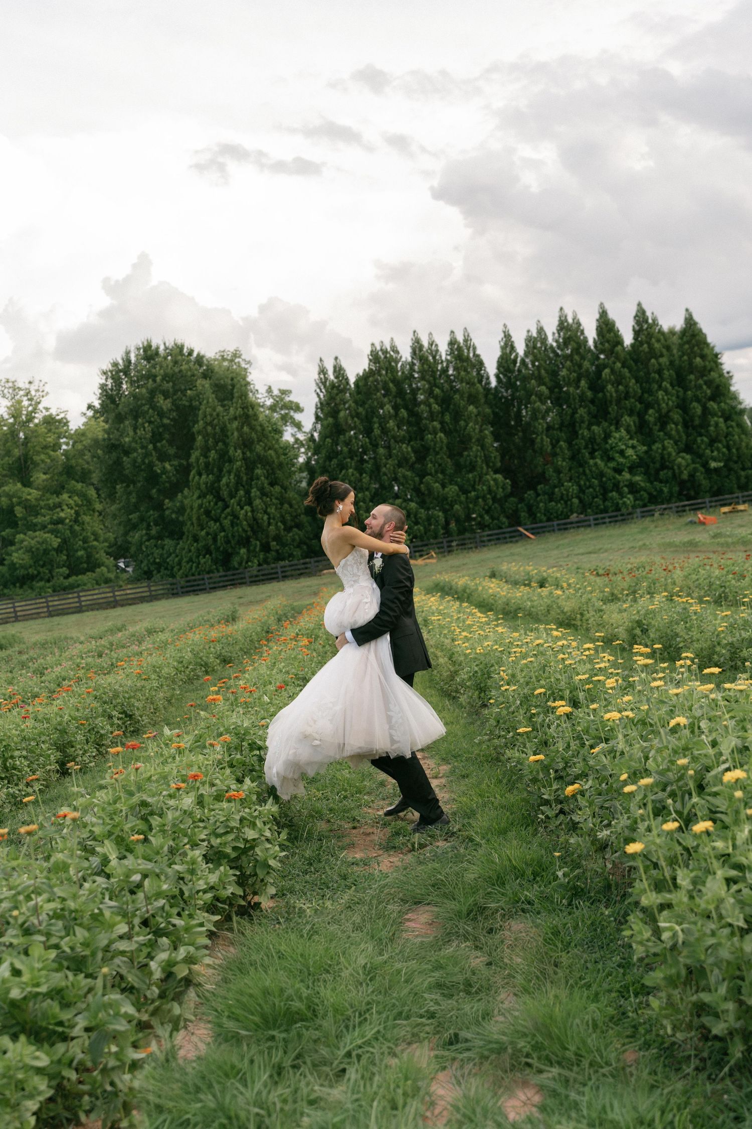 Groom lifting bride in white gown in a lush green flower field, surrounded by tall trees on their wedding day.