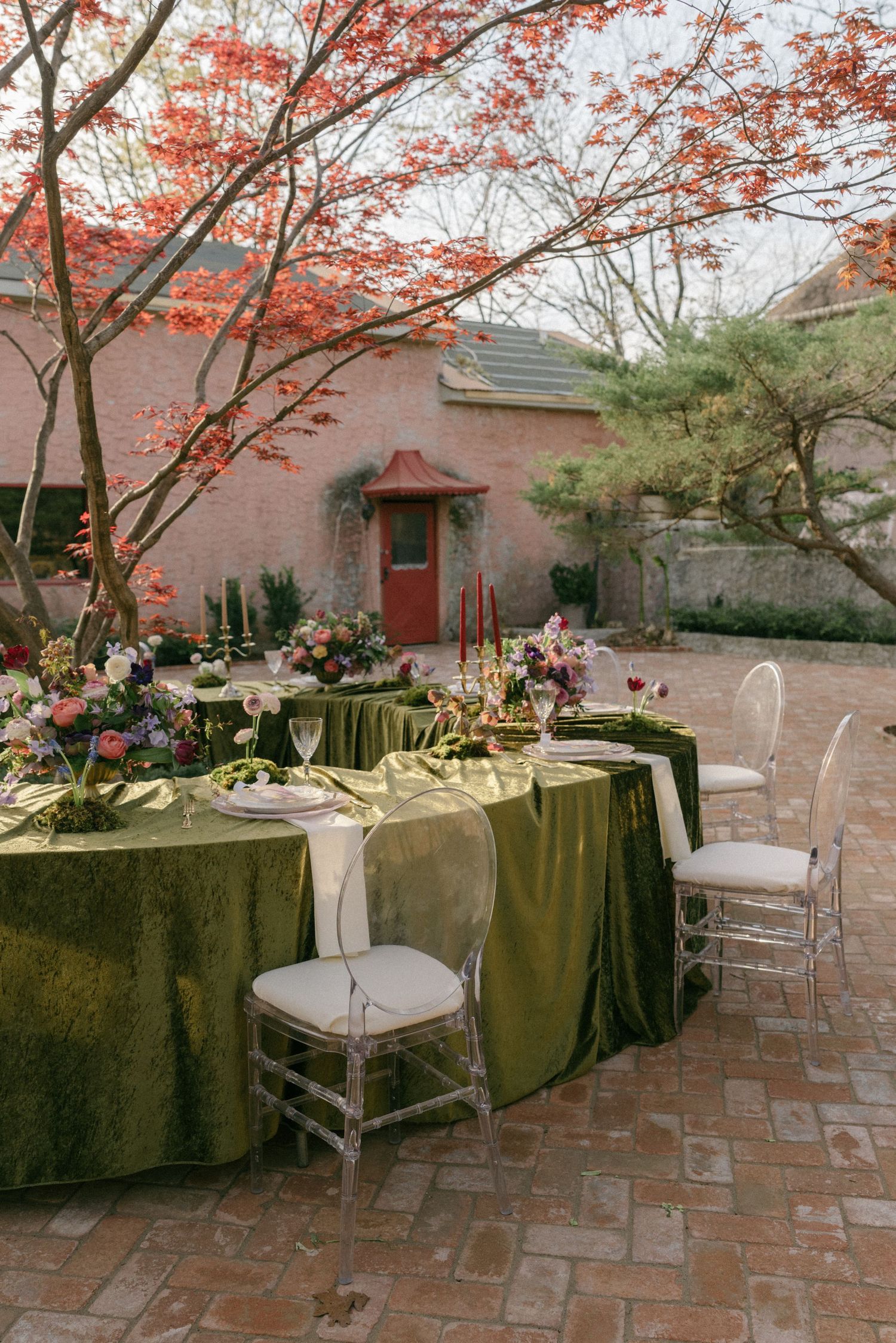 Elegant outdoor wedding reception table with olive green velvet linens, colorful floral arrangements, and ghost chairs on a brick patio.
