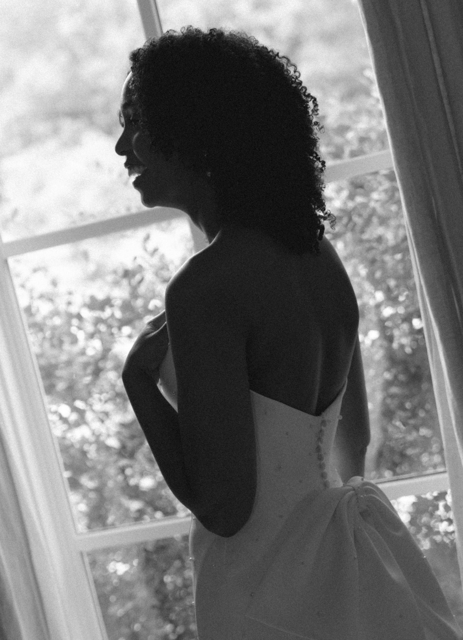 Black and white photo of a bride in a strapless wedding gown standing by a window.