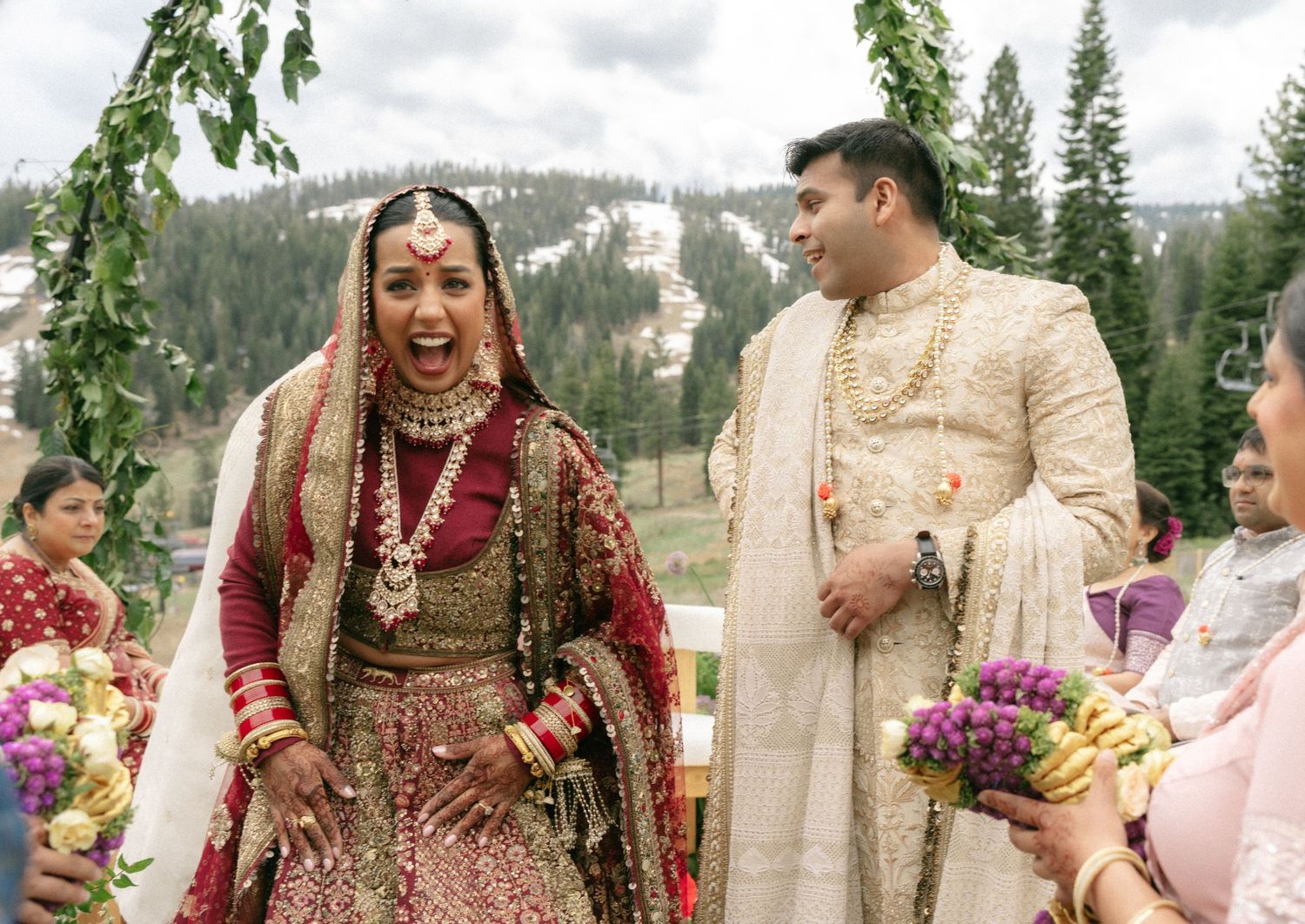 Joyful Indian bride in red lehenga laughs with groom in cream sherwani at outdoor mountain wedding ceremony.