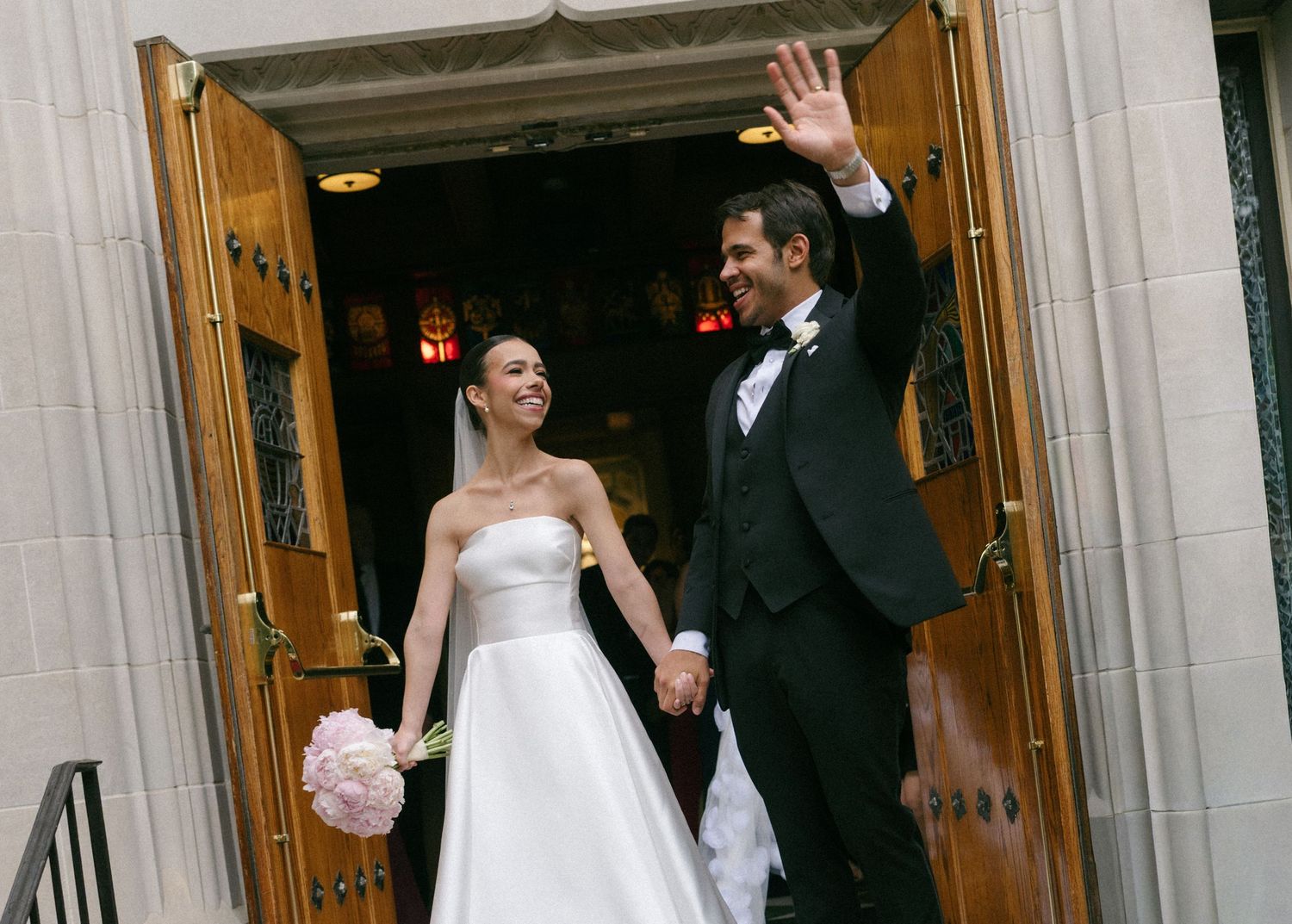 Newlywed couple holding hands and waving as they exit a church after their wedding ceremony.