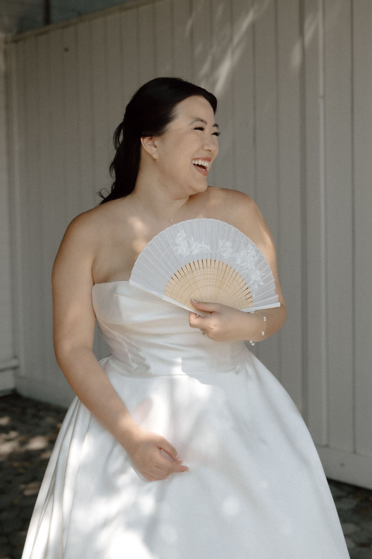 Laughing bride in strapless white gown holding decorative white hand fan outdoors on sunny wedding day.