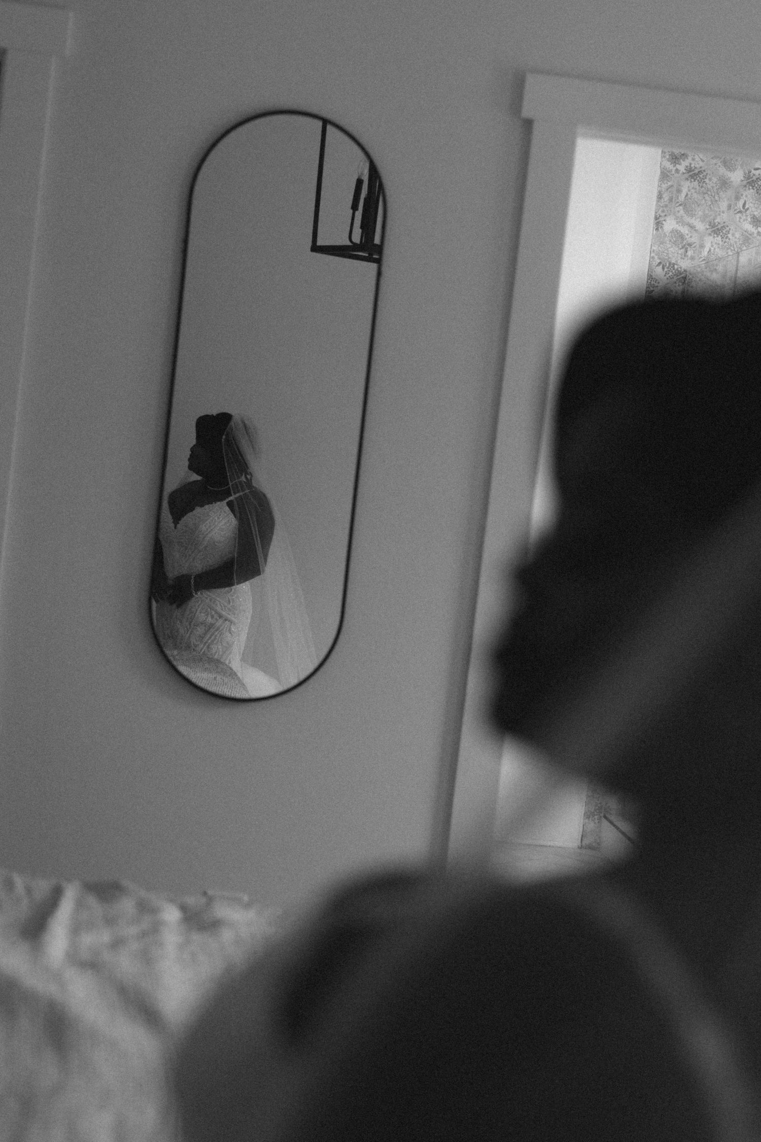 Black and white photo of a bride reflected in an oval mirror, holding her bouquet in a softly lit room.