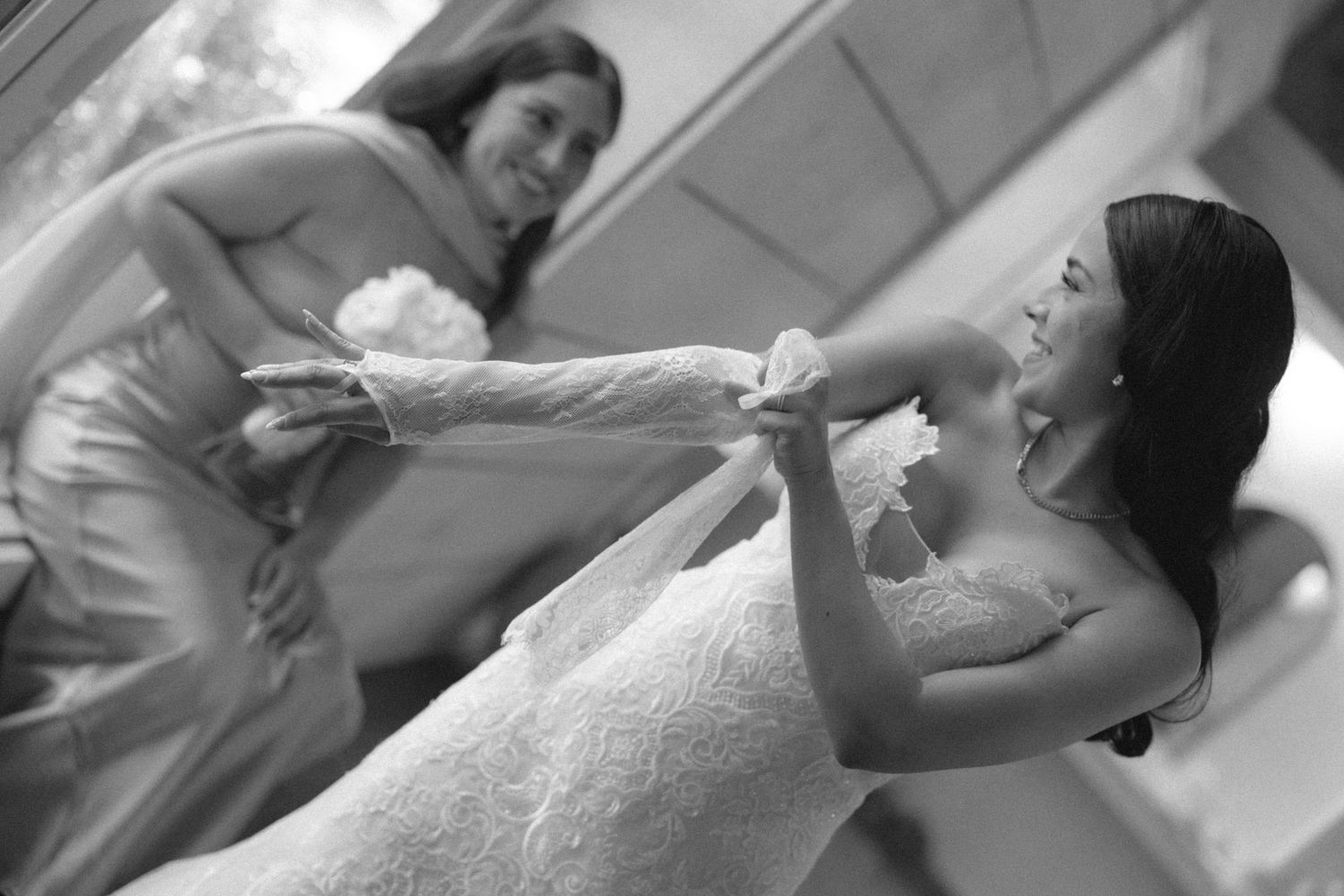 Black and white photo of a bride in a lace gown having her veil adjusted by a bridesmaid in a satin dress.