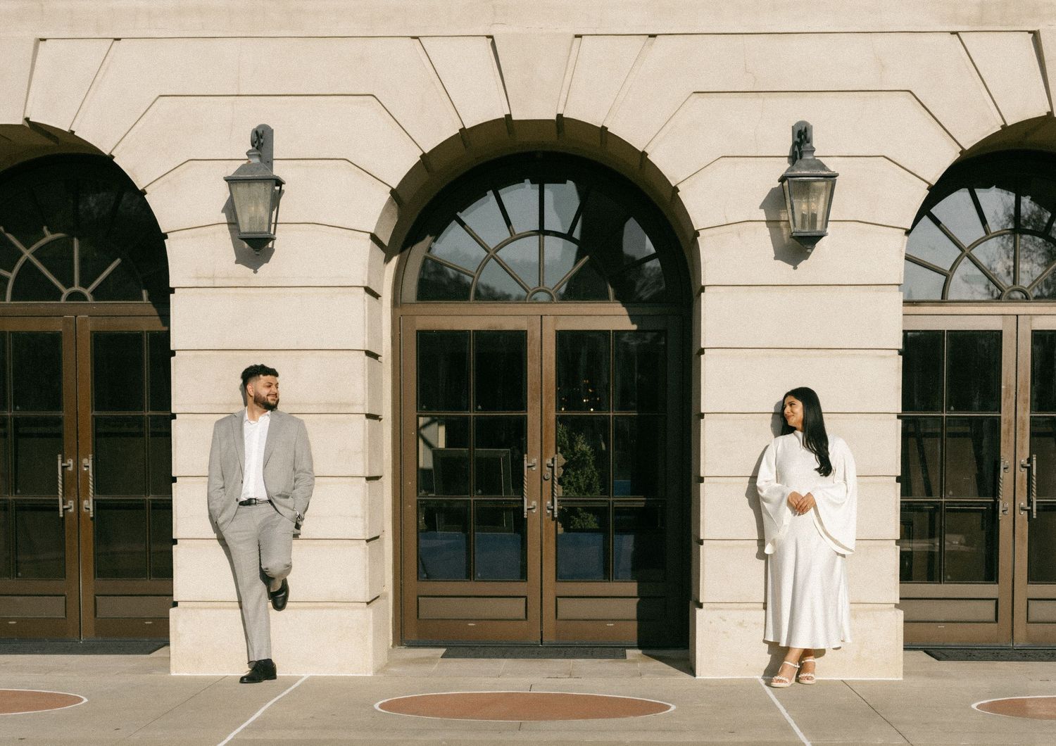 Couple posing apart in front of elegant beige arched building with gold doors and wall sconces.