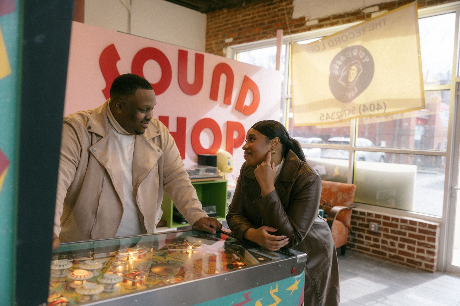 Two people laugh together at a pinball machine inside a colorful Sound Shop music store.