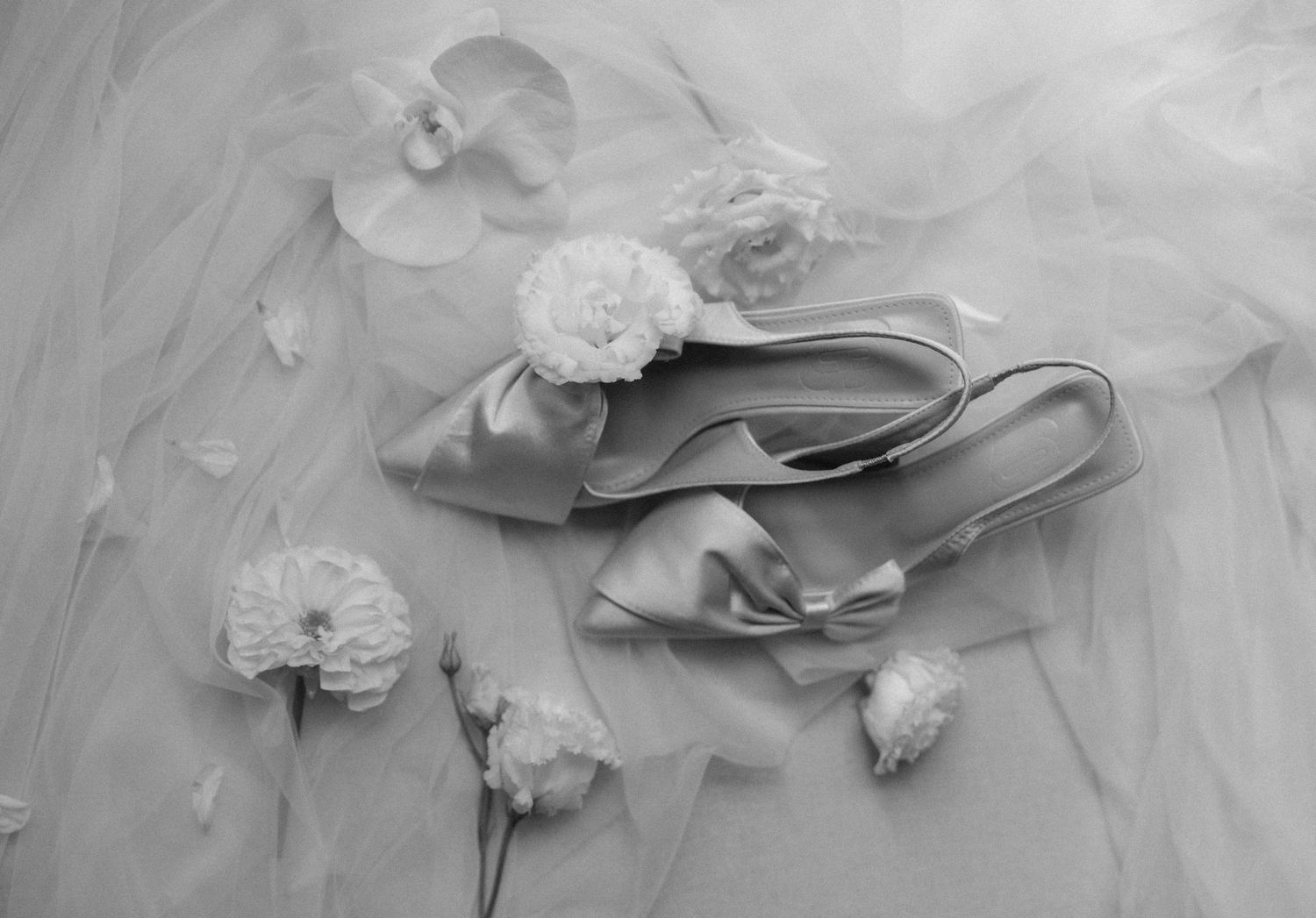 Elegant bridal satin heels with bow detail surrounded by white flowers on soft tulle fabric in black and white.