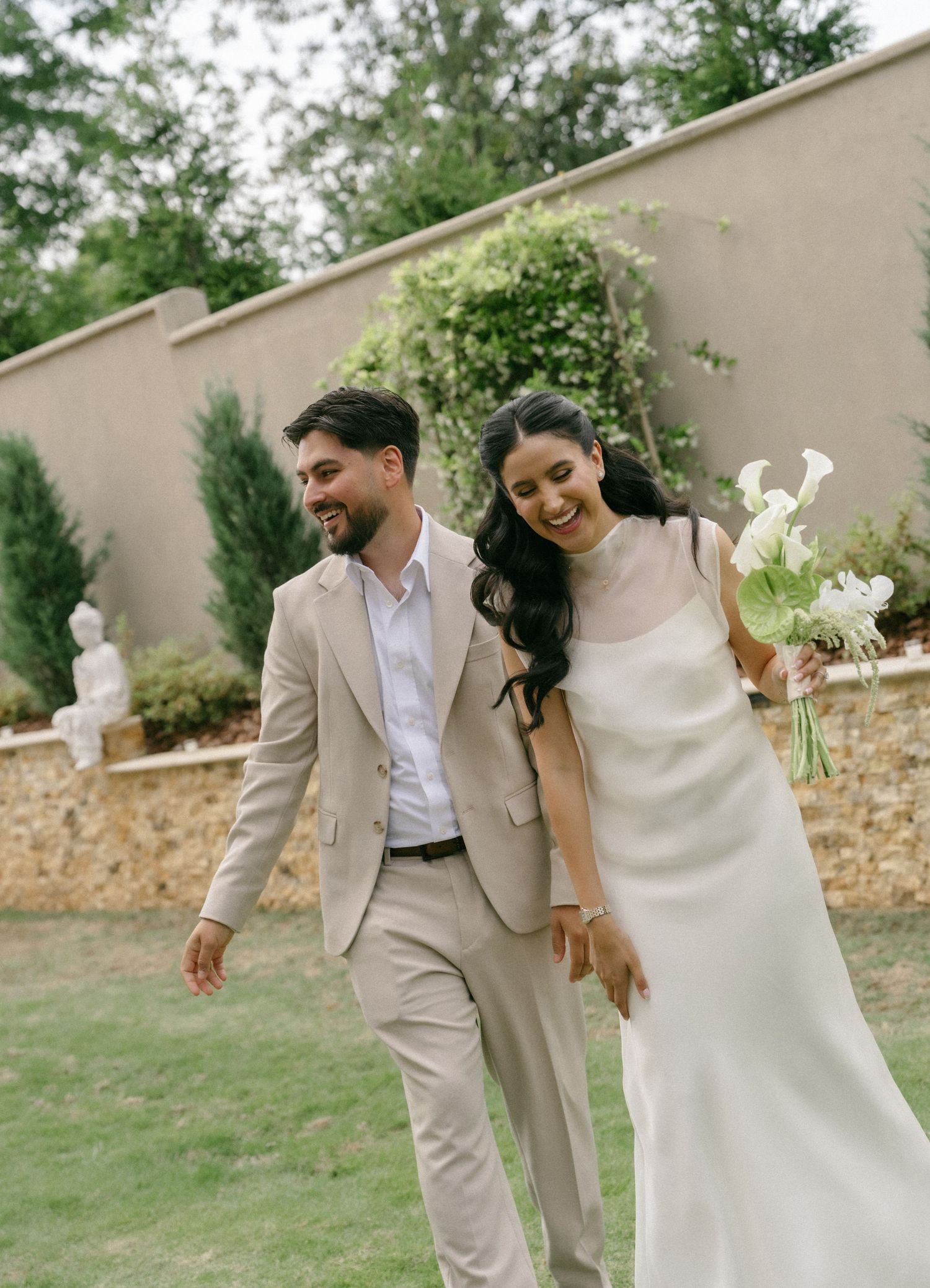 A smiling bride in a white gown holding calla lilies walks with a groom in a beige suit at an outdoor wedding.