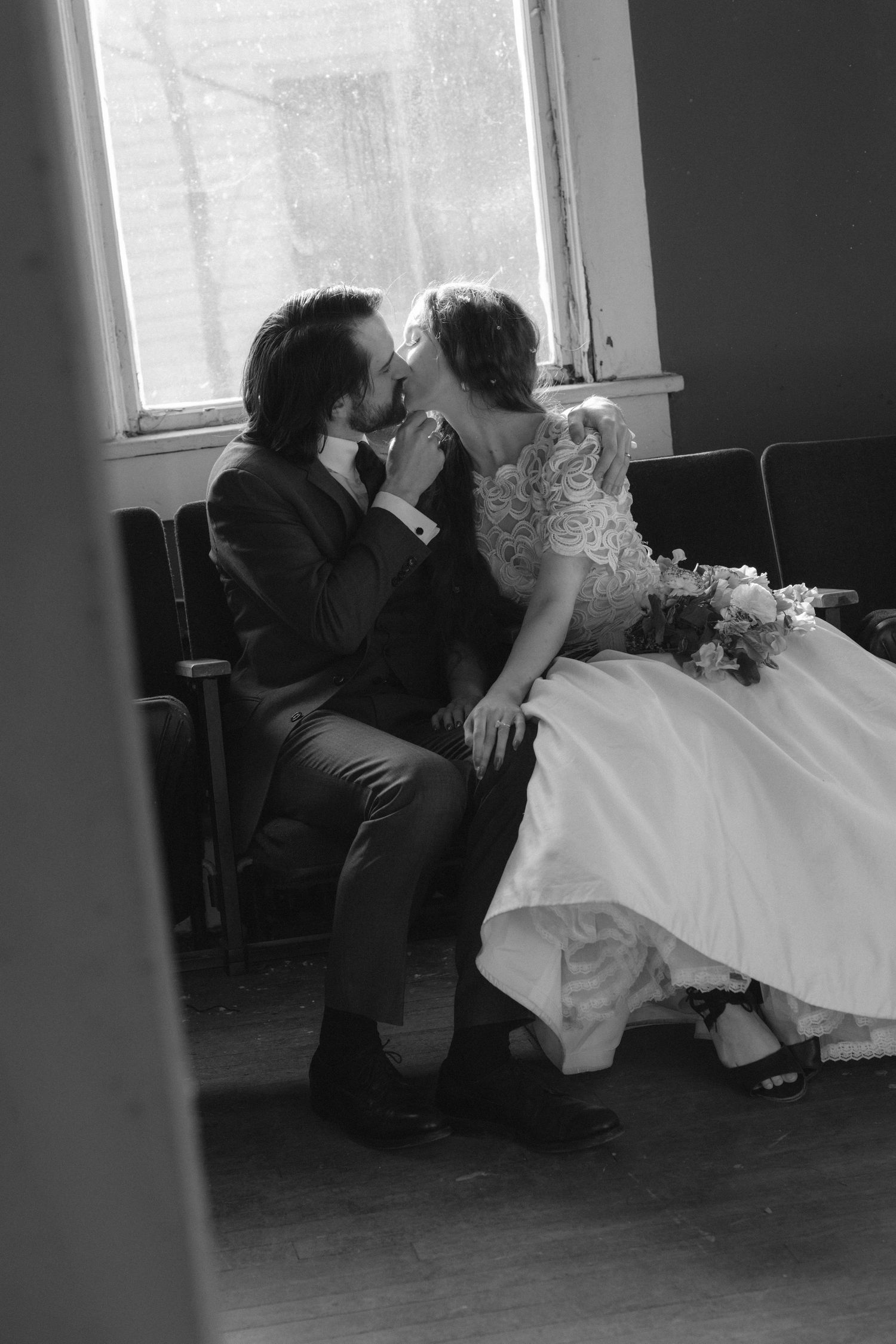 Black and white photo of a bride and groom sharing a kiss while seated near a window on their wedding day.