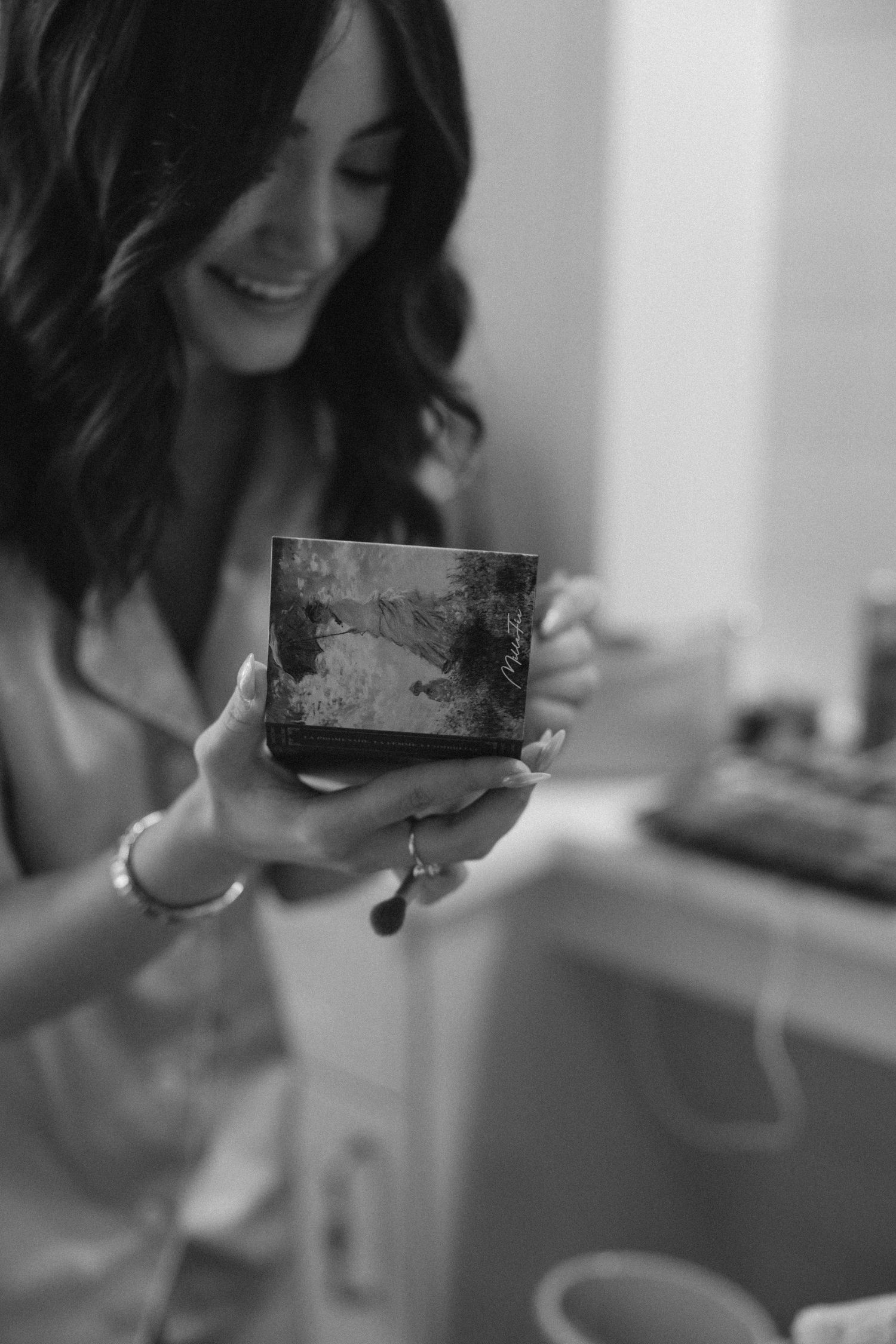 Black and white photo of smiling brunette woman holding a small decorative box in a bathroom.
