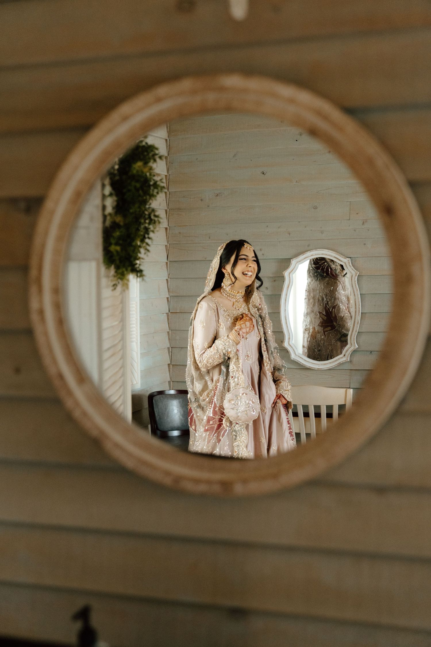 Woman in elegant floral dress smiling, reflected in a round wooden mirror inside a rustic wood-paneled room.