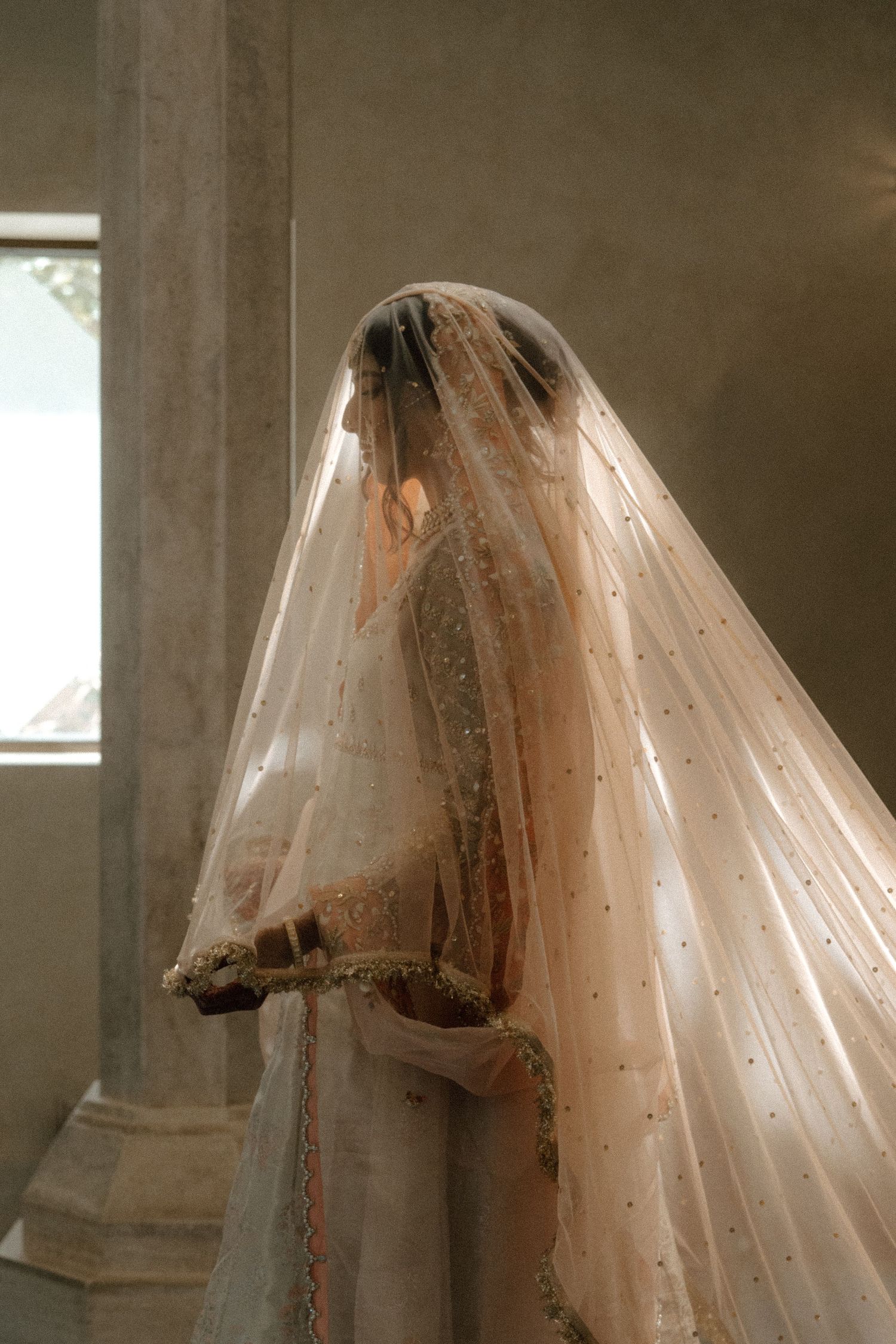 Bride in ornate South Asian wedding attire with embroidered veil, standing in soft natural light.