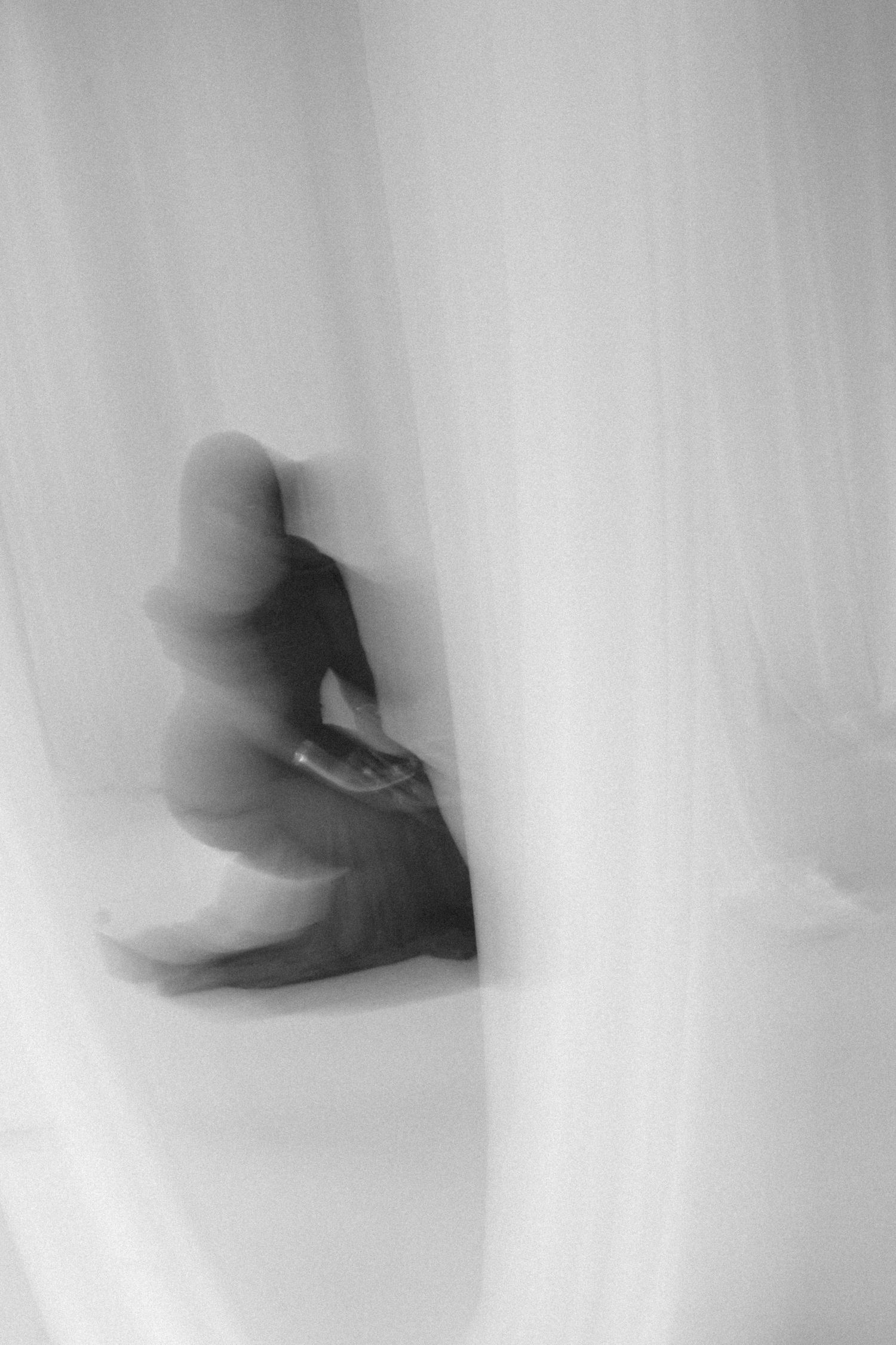 Blurry black and white photo of a person sitting behind sheer white curtains, creating a mysterious silhouette effect.