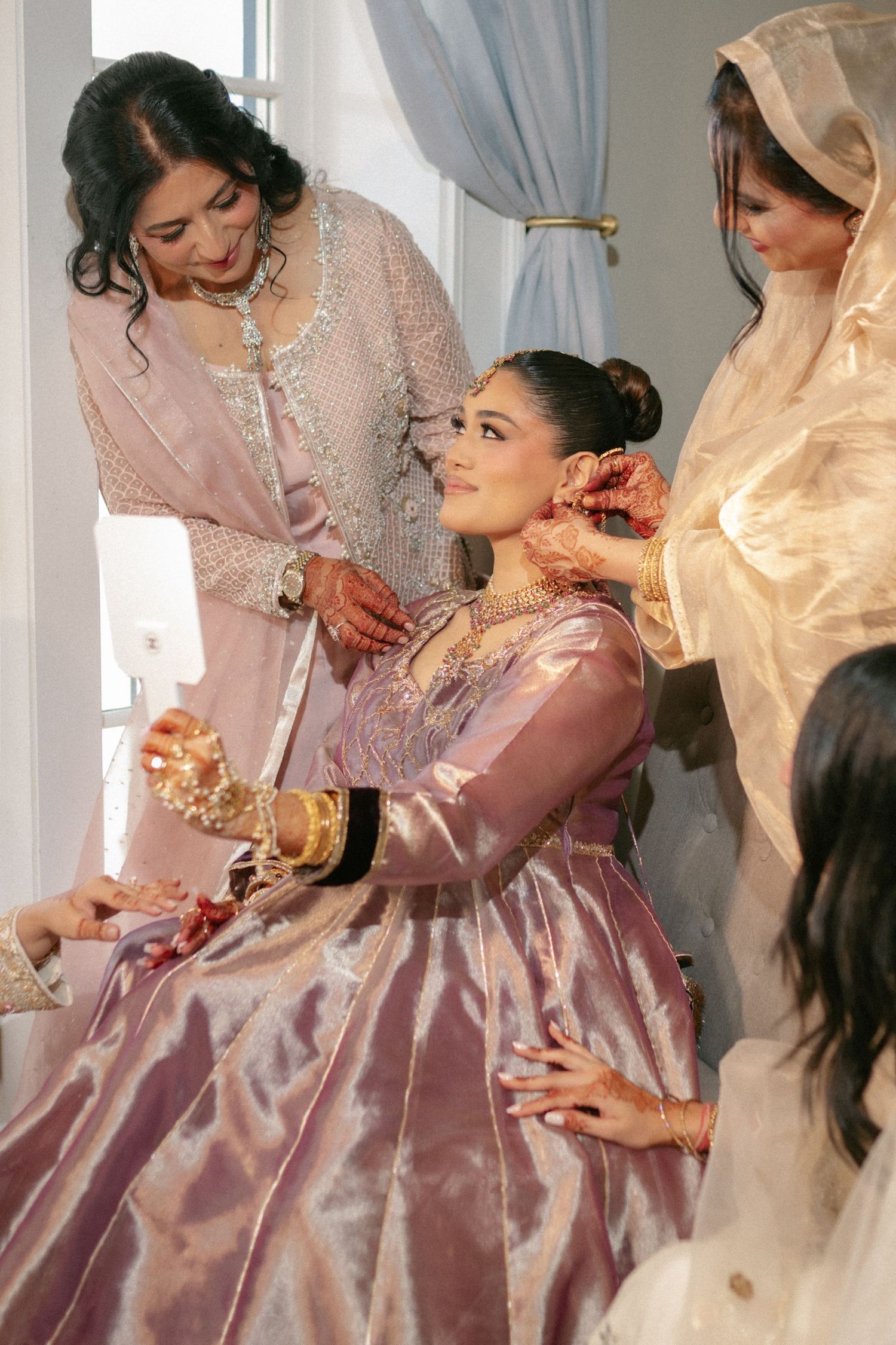 A bride in a pink lehenga sits as women in traditional South Asian attire help her prepare for her wedding ceremony.