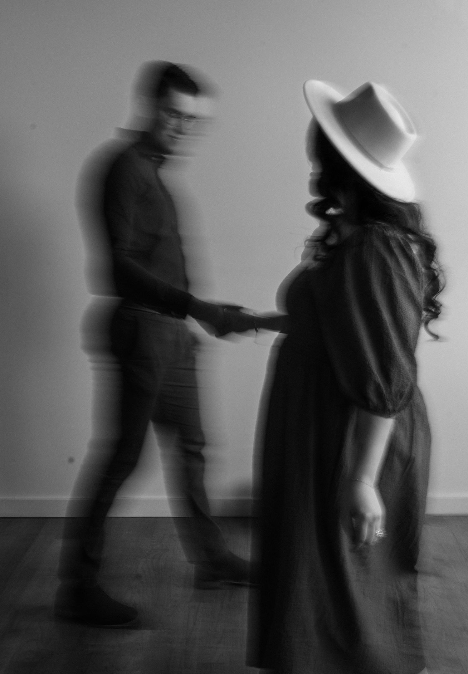 Black and white motion blur photo of a pregnant woman in a hat holding hands with a man.