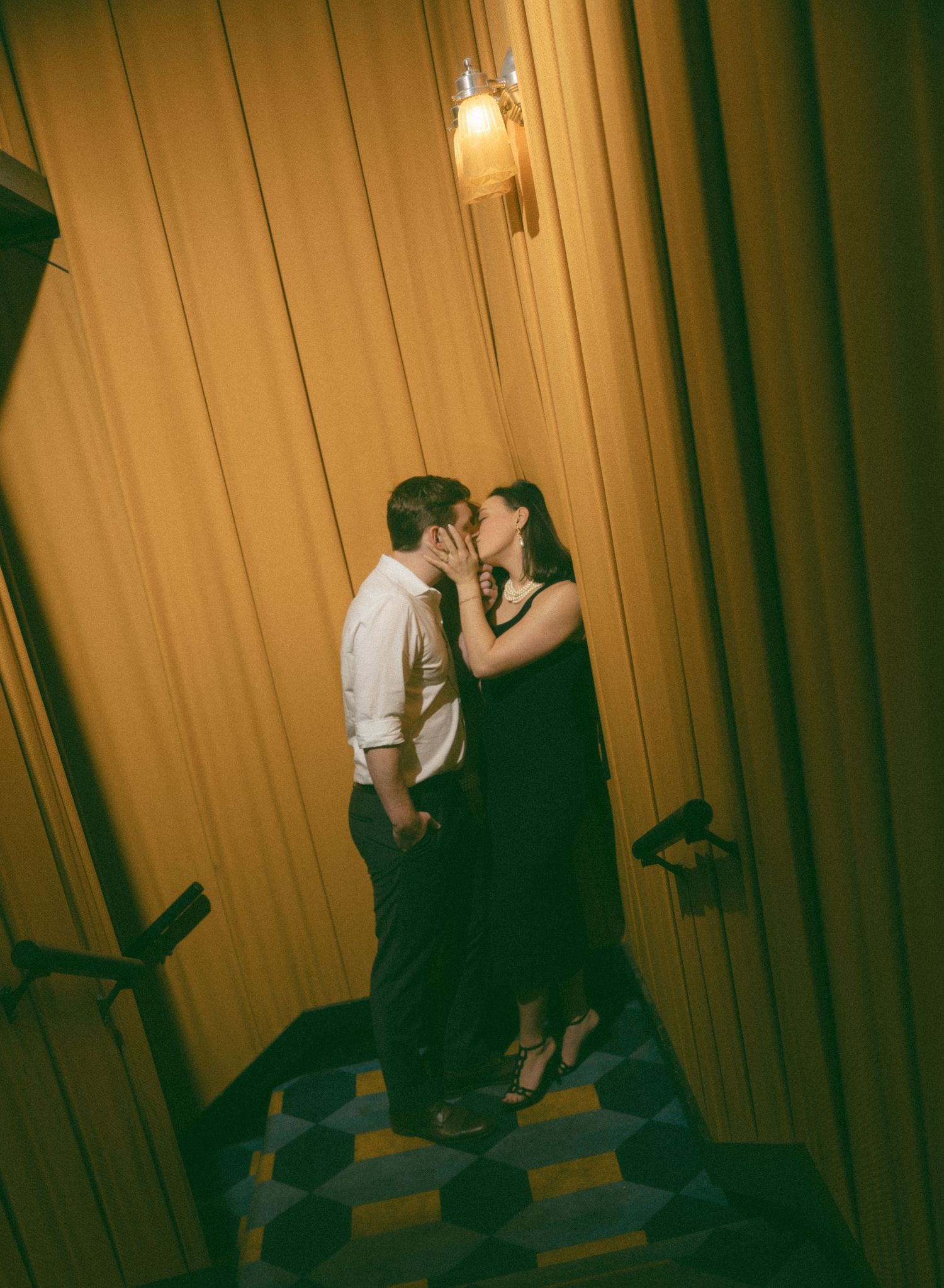 Couple sharing a romantic kiss in a warmly lit corridor with golden curtains and a decorative wall sconce above.